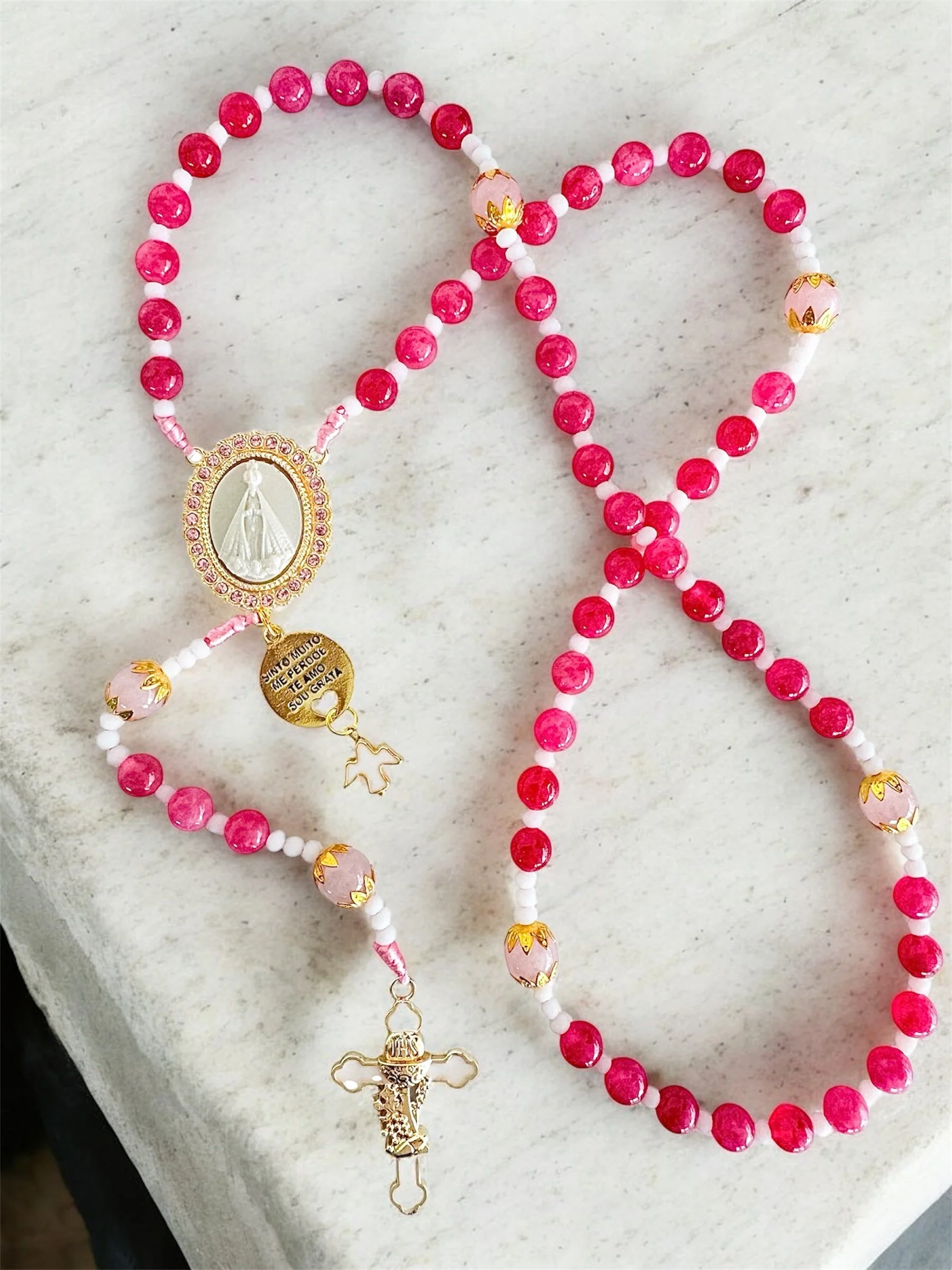 A religious rosary necklace with pink beads, gold accents, a gold cross, and a medallion with a religious figure, resting on white fabric.