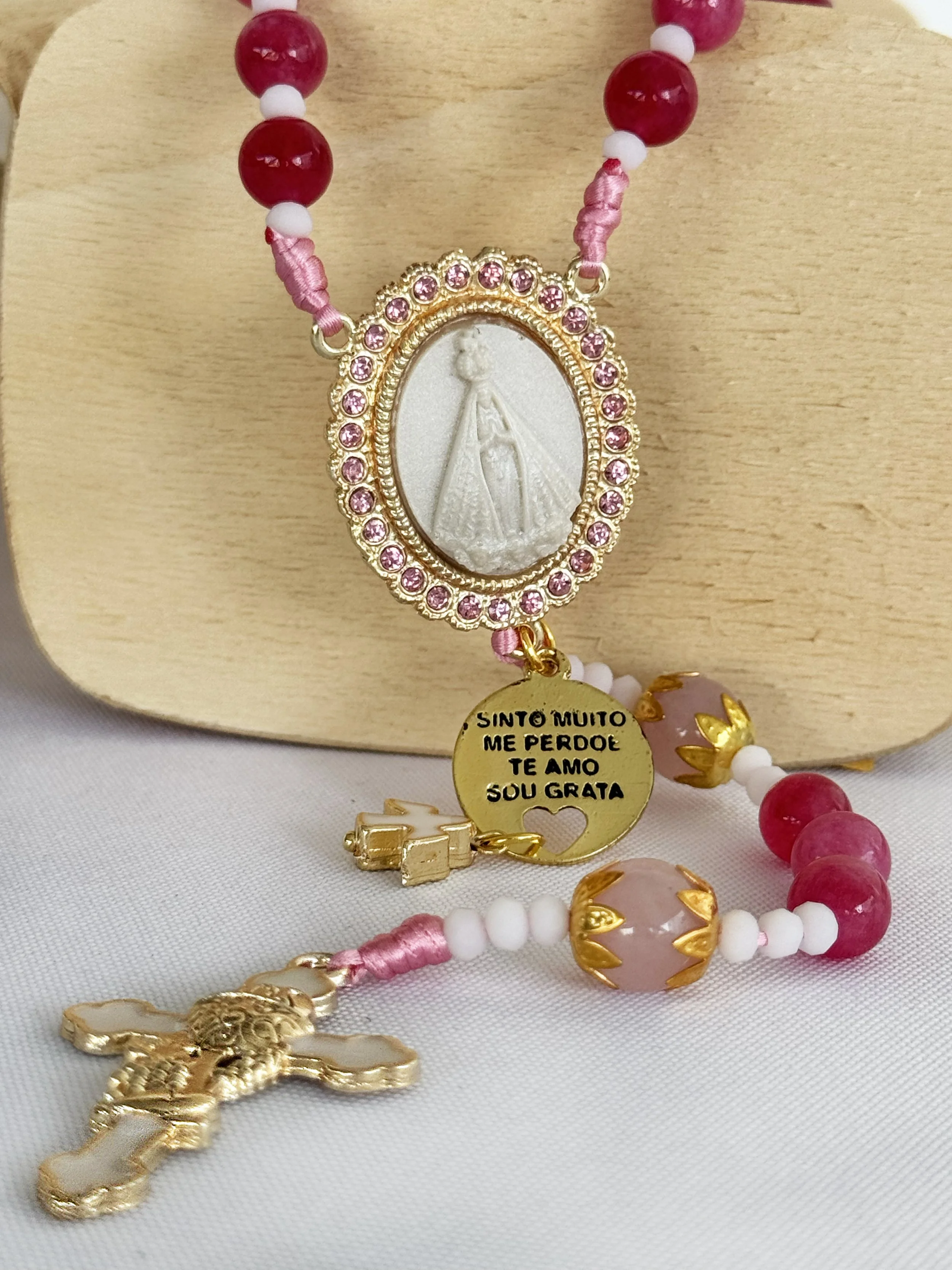 Close-up of a religious necklace with a white oval pendant featuring an embossed image of the Virgin Mary, surrounded by pink rhinestones. The necklace has pink, white, and red beads with gold accents, and a gold cross pendant at the bottom.