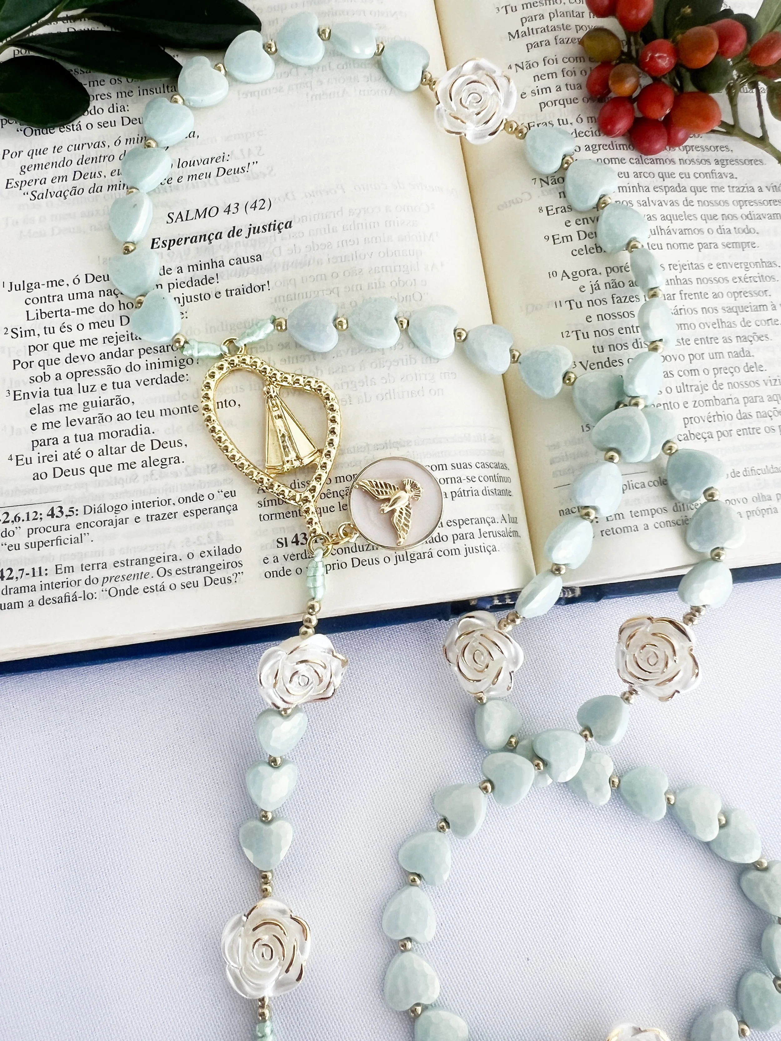 An open book with a blue beaded necklace, a ring with a hummingbird design, and a gold heart-shaped peace symbol pendant laid on top, with a bunch of red berries and green leaves at the top right corner.