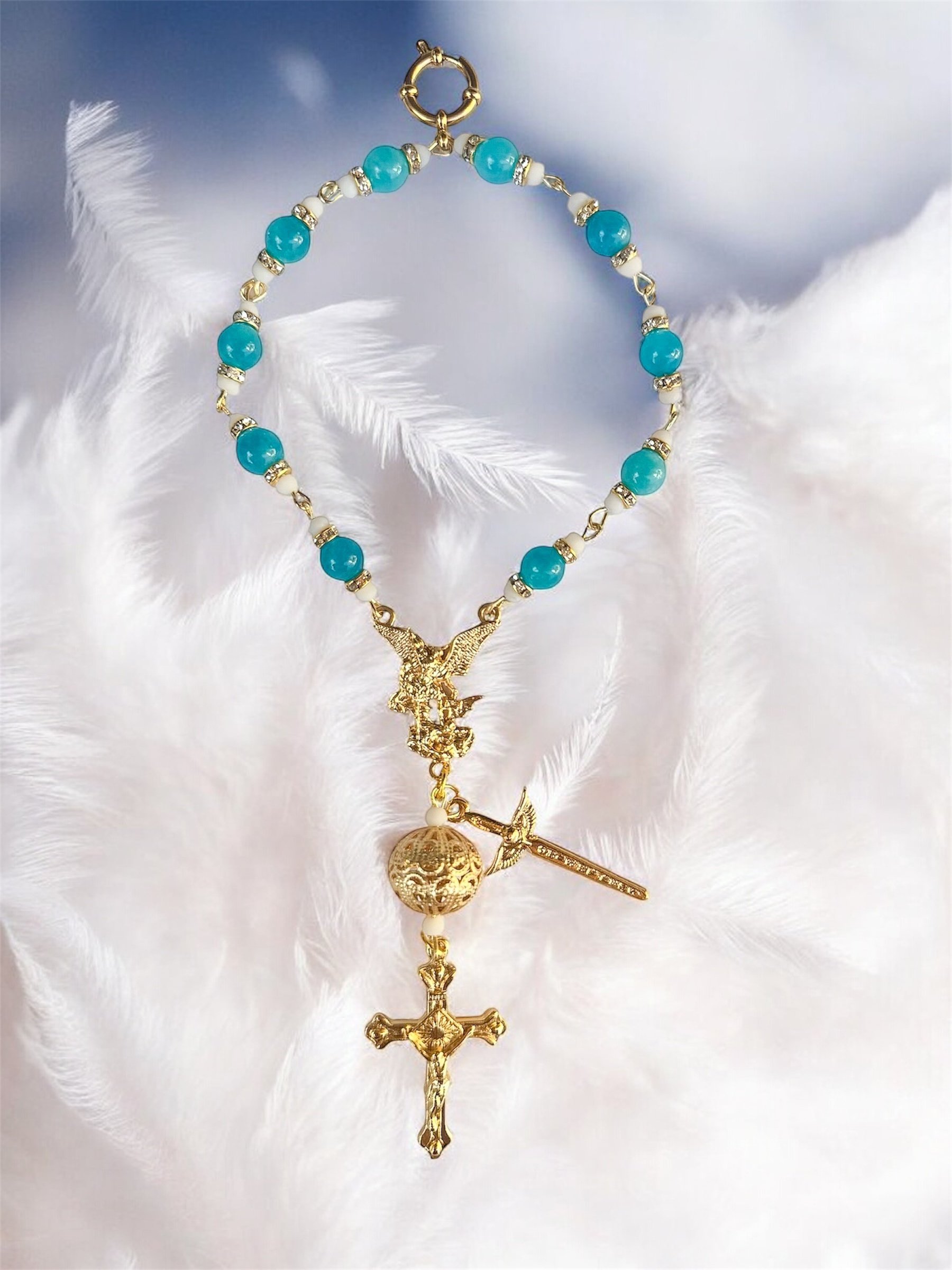 A gold rosary with blue beads, white accent beads, and a gold crucifix, resting on white feathers with a blurred background.
