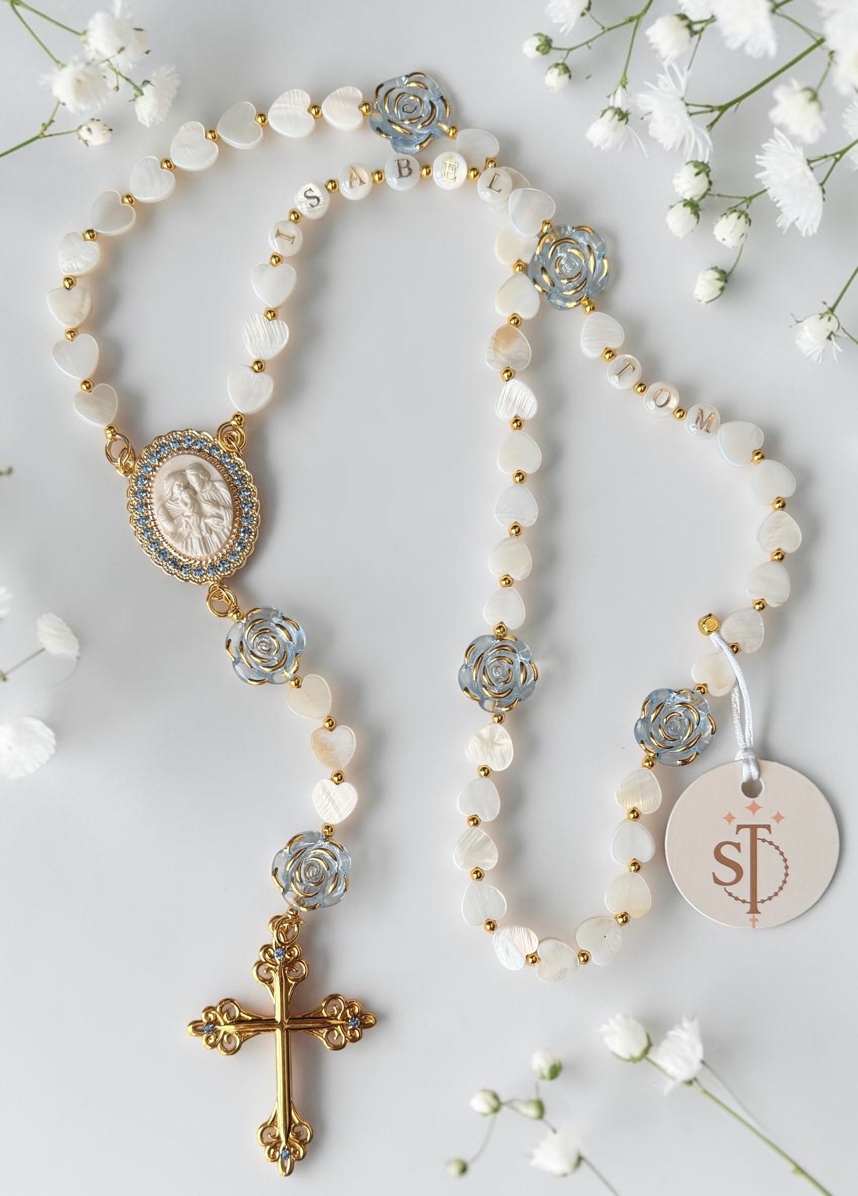 Religious rosary beads with a crucifix and a medal of the Virgin Mary, decorated with small heart-shaped white beads, round white beads with letter engravings spelling 'SABEL' and 'LOVO', silver roses, gold accents, and white flowers scattered around