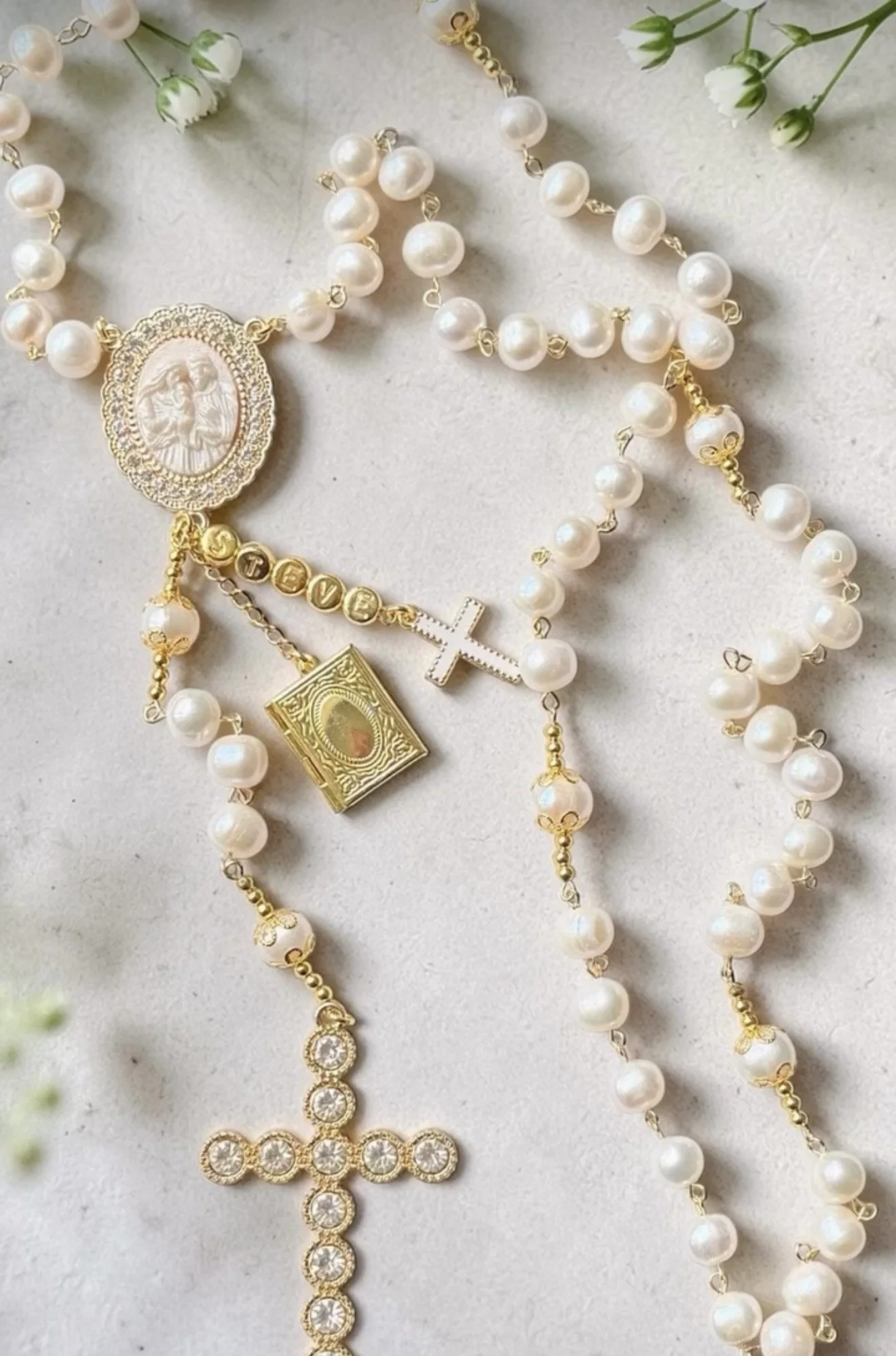 Multiple pearl necklaces with gold and silver charms, including a crucifix, a cameo, a cross with the word 'Jesus', a small gold book, and a gold chain with circle rhinestones, arranged on a white surface with small white flowers nearby.