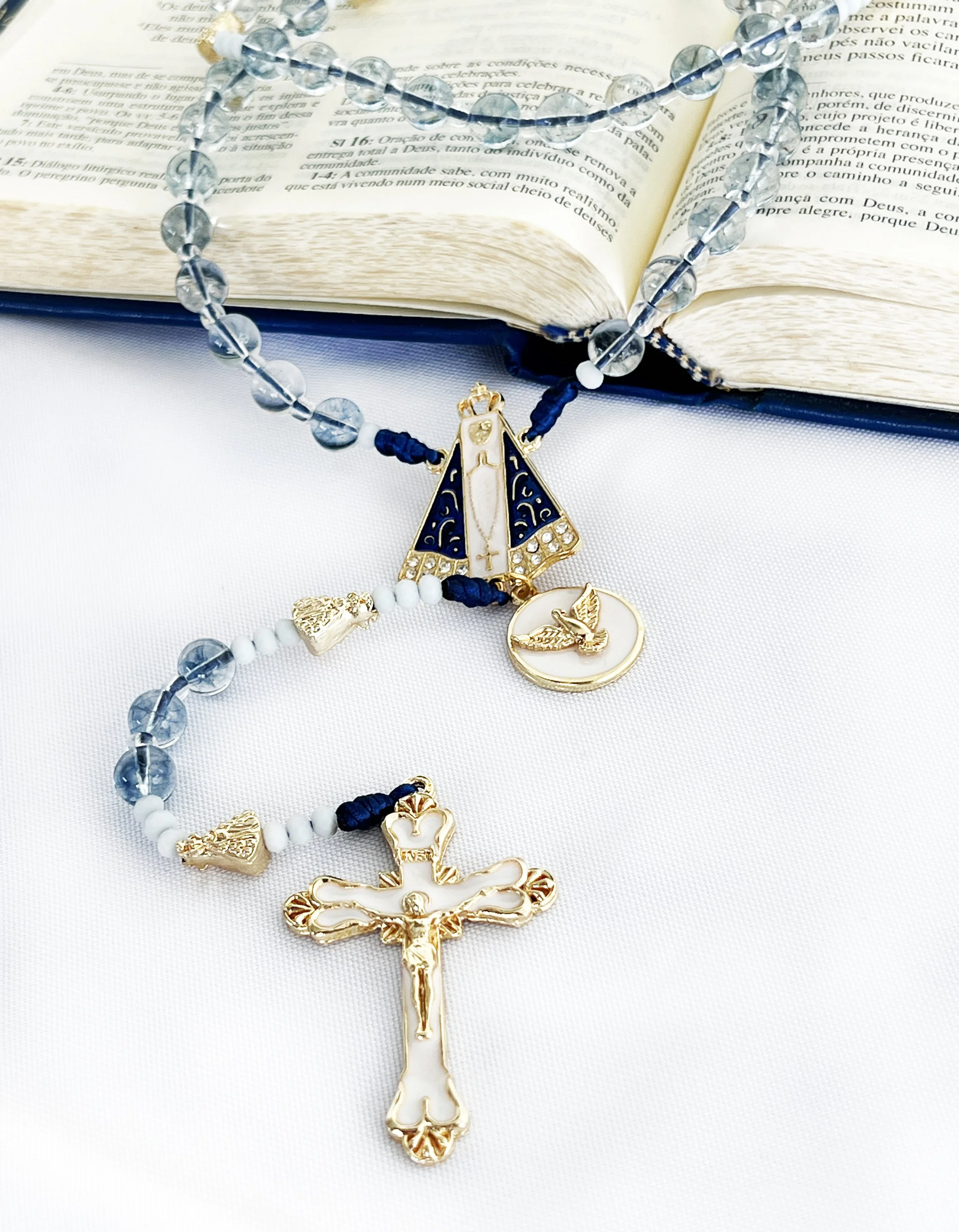 A gold and white crucifix necklace with a dove in the center, resting on an open book with text, and a string of blue and clear beads draped over the book.