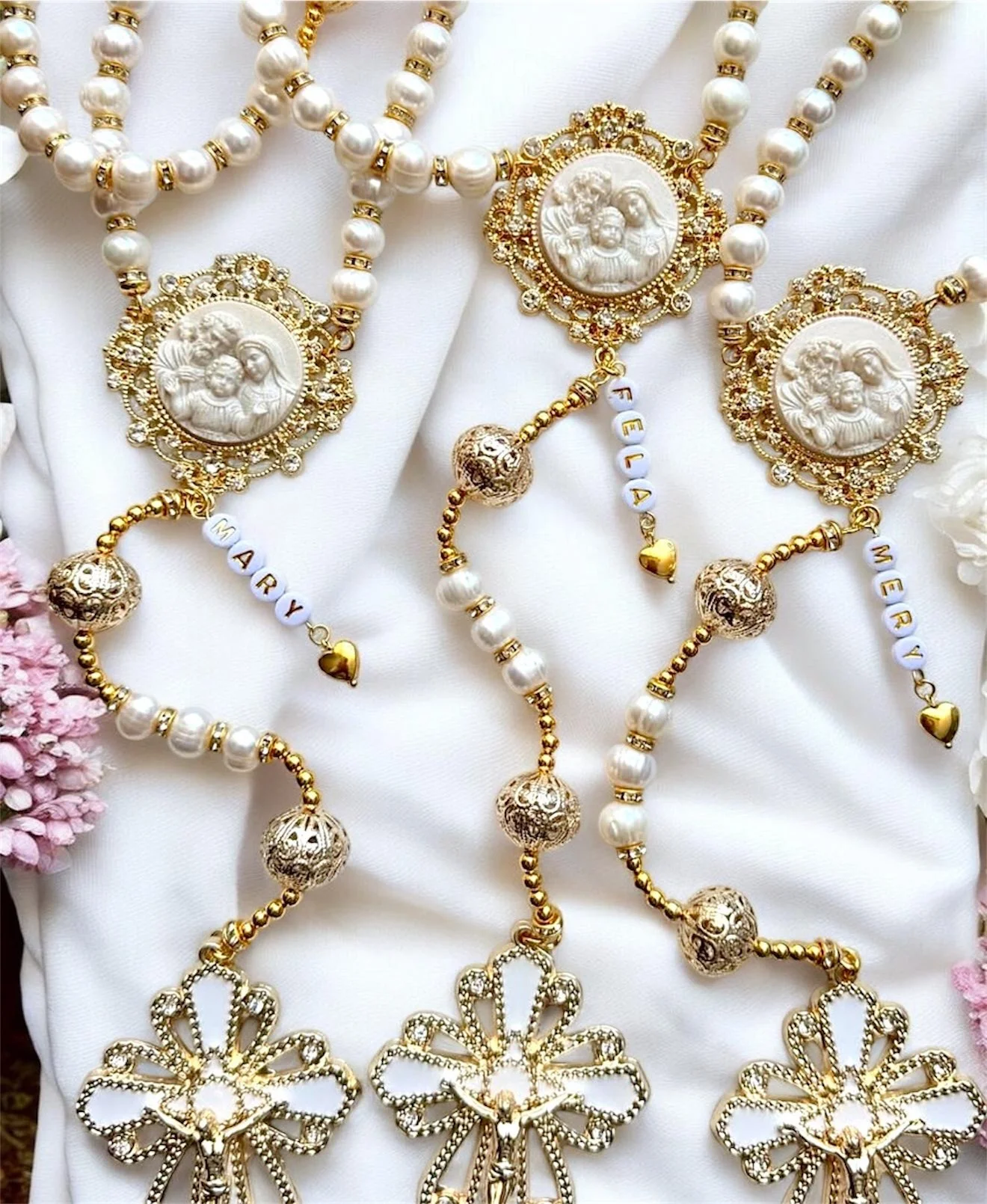 Multiple pearl necklaces and bracelets with gold medallions and charms, some inscribed with names, set on a white fabric background.