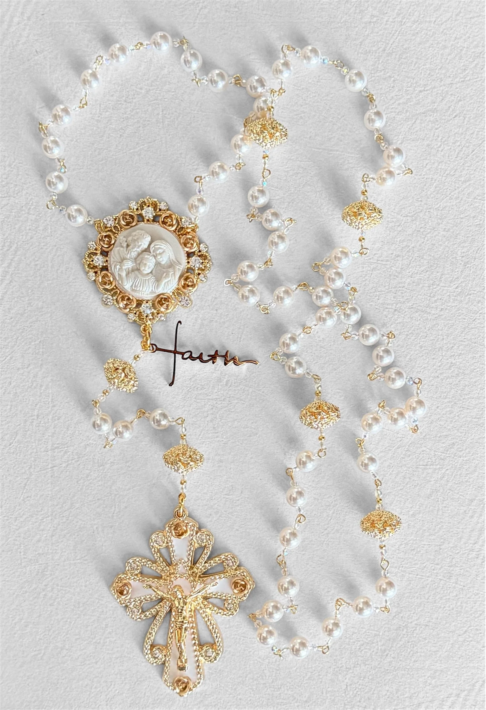 Gold and pearl religious rosary with a crucifix, featuring a central medallion depicting a religious scene and a gold cross with a bow at the bottom, all laid out on a light gray surface.