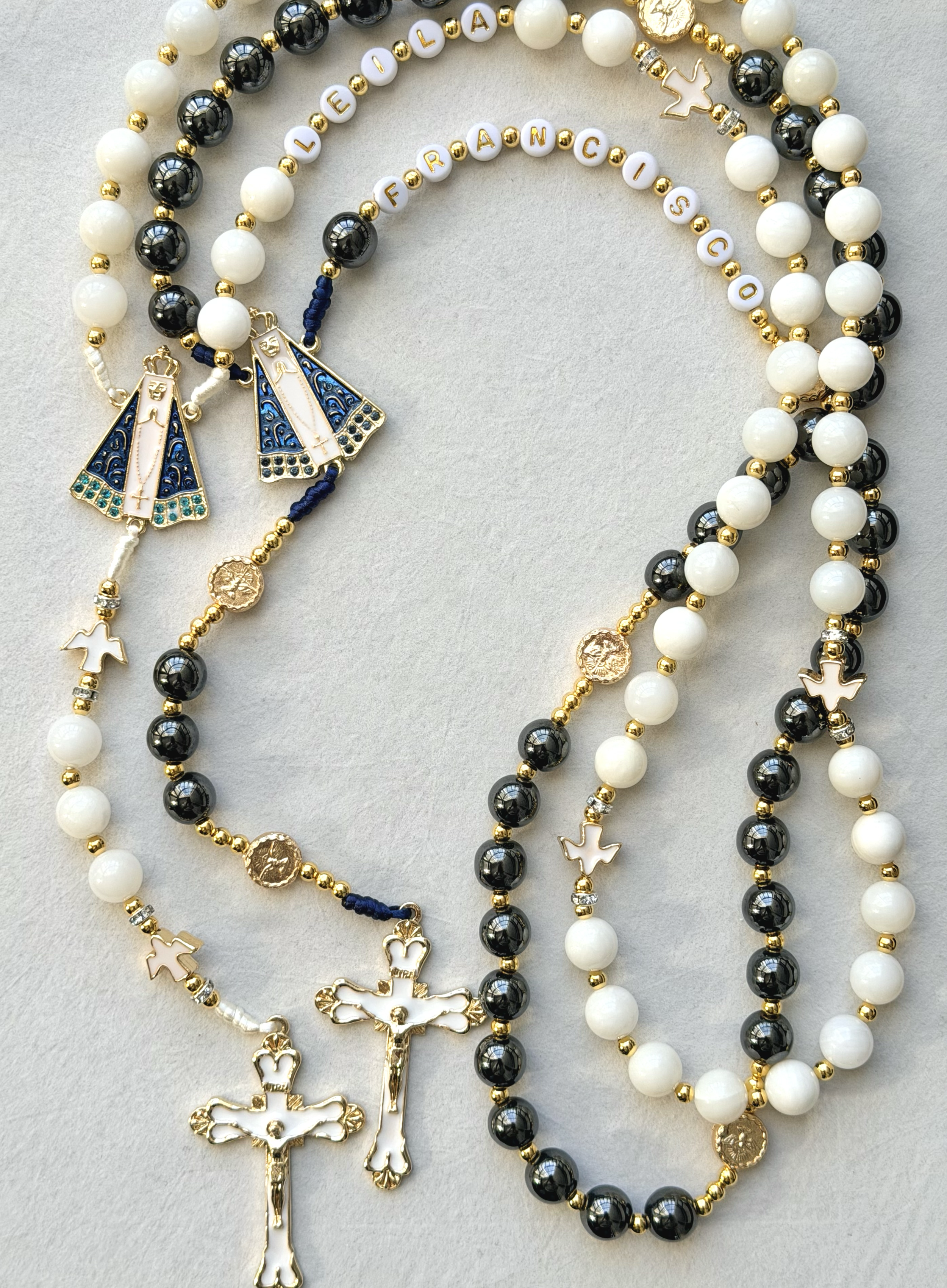 Collection of religious beaded necklaces and rosaries with gold, black, and white beads, featuring crosses and religious pendants, arranged on a light surface.