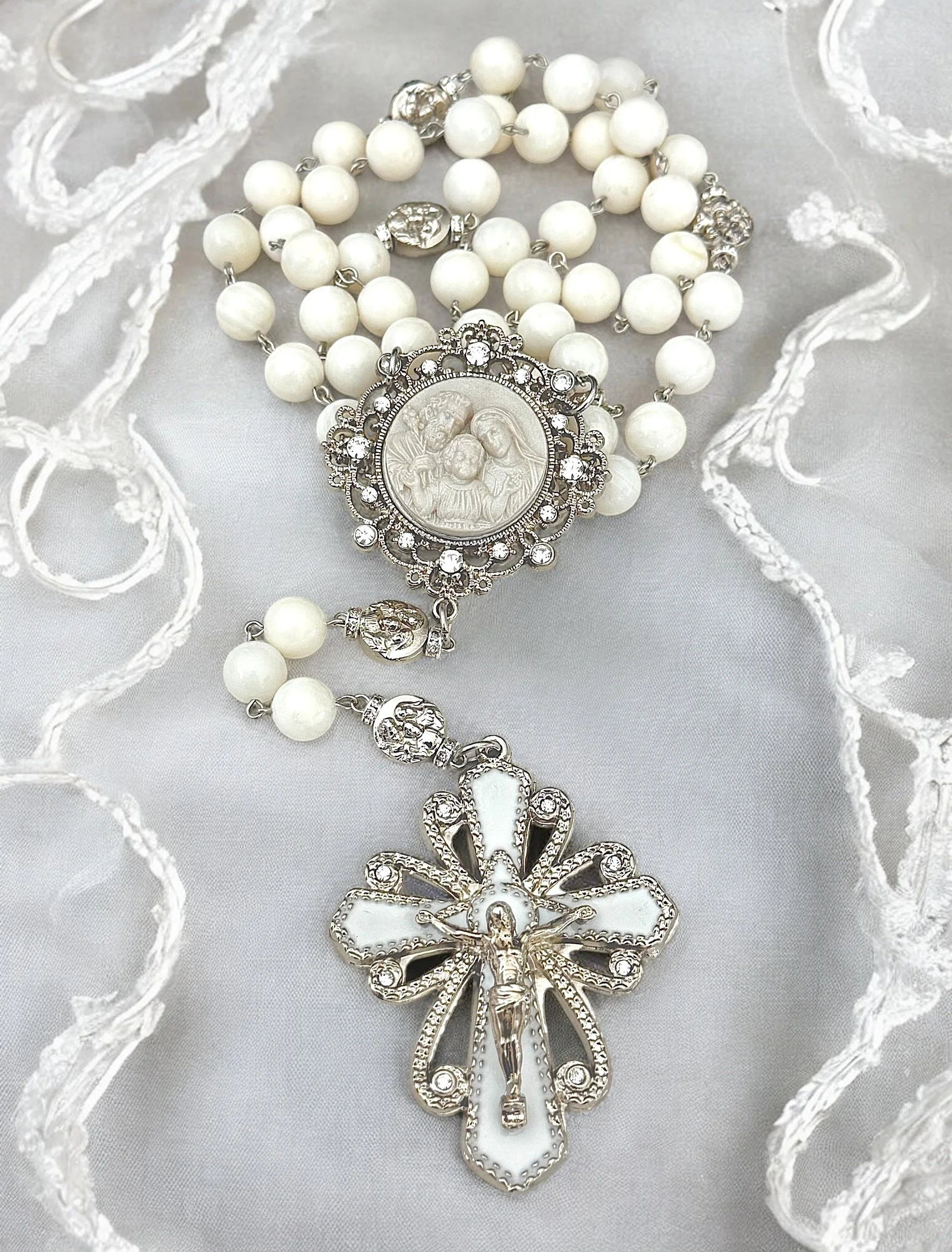 A pearl necklace with a silver religious pendant and decorative charms, displayed on white lace fabric.
