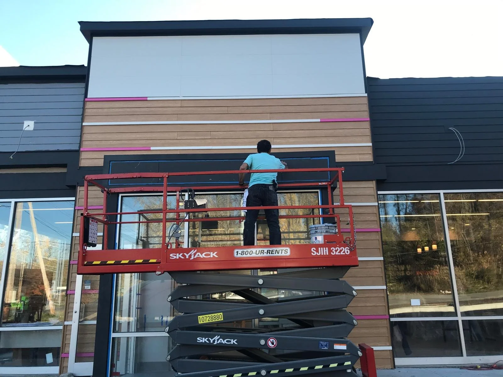A person on a red scissor lift working on the exterior of a building, which has a wood and black panel facade with large windows.