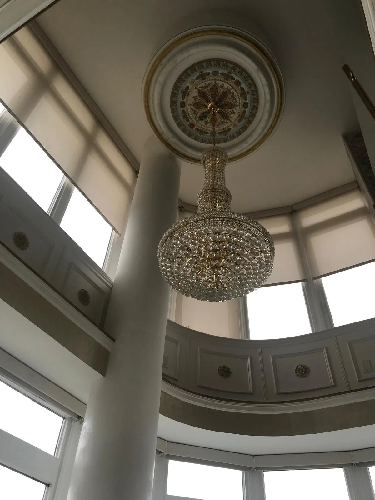 Elegant chandelier hanging from a circular ceiling medallion in a grand interior space with tall windows and decorative molding.