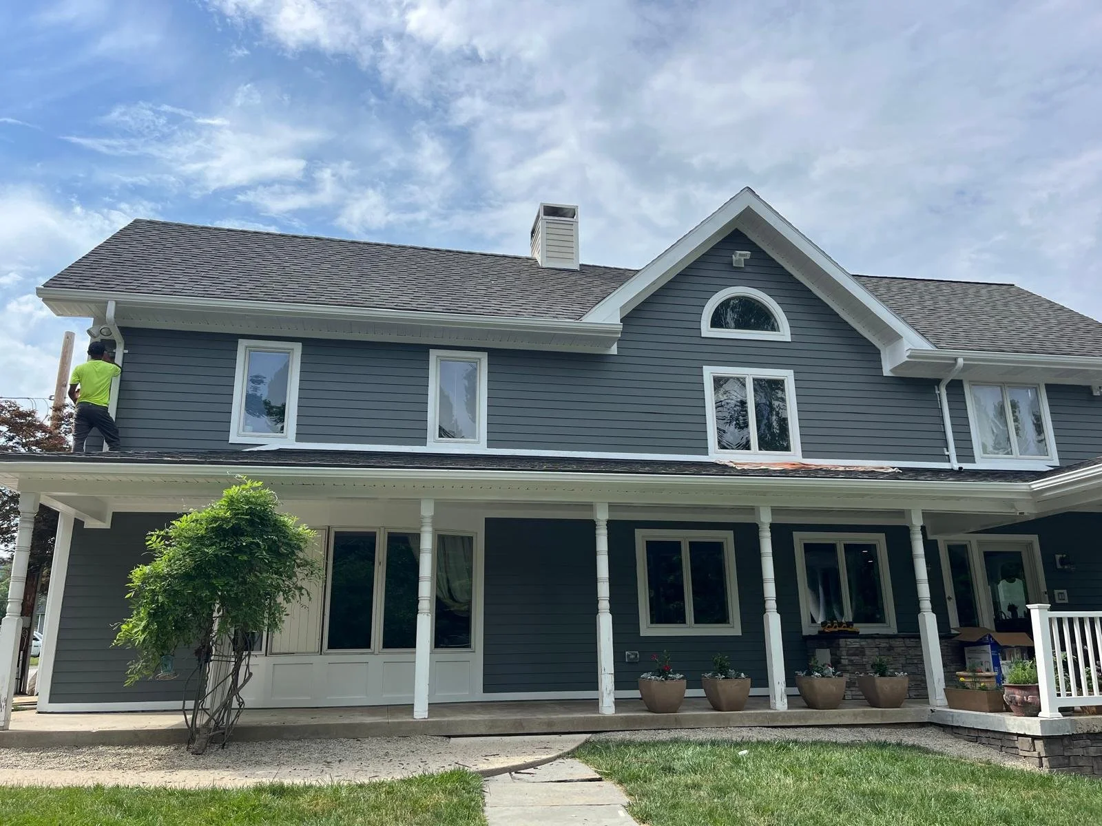 A large two-story house with gray siding, multiple windows, and a porch with white pillars. A person in a neon green shirt is working on the roof. There are potted plants in front, and the sky is partly cloudy.