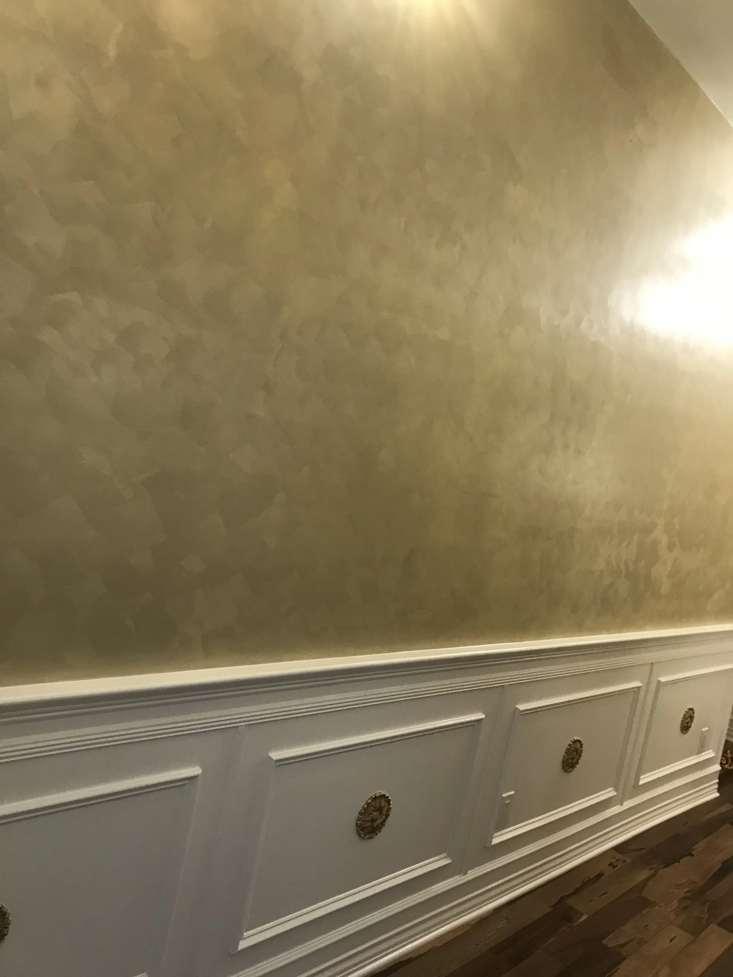 A white dresser with gold-colored decorative handles and ornate detailing at the bottom, set against a light-colored, textured wall with molding at the bottom.