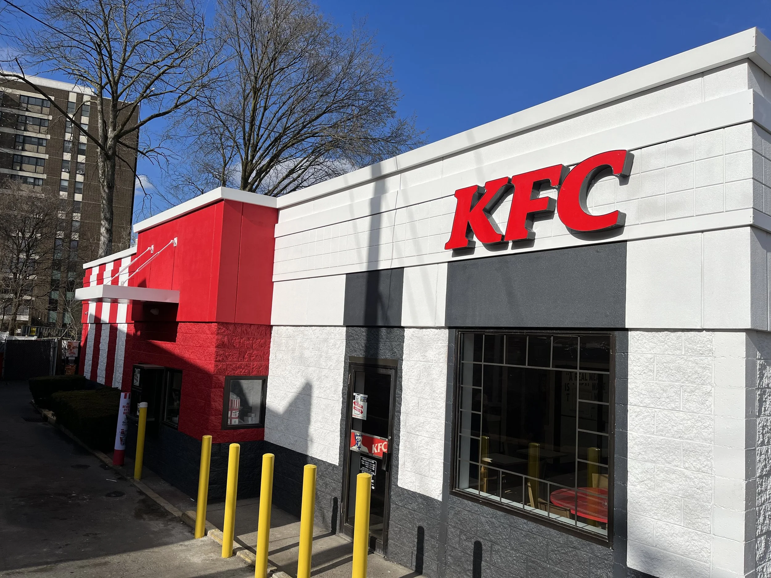 Exterior of a KFC restaurant with white and red decor, large red letters spelling 'KFC', yellow safety bollards, leafless trees, and a clear blue sky in an urban area.