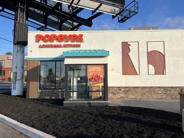 Exterior of a Popeyes Louisiana Kitchen restaurant with a glass entrance, a small promotional sign, and modern decorative wall art, under a clear blue sky.