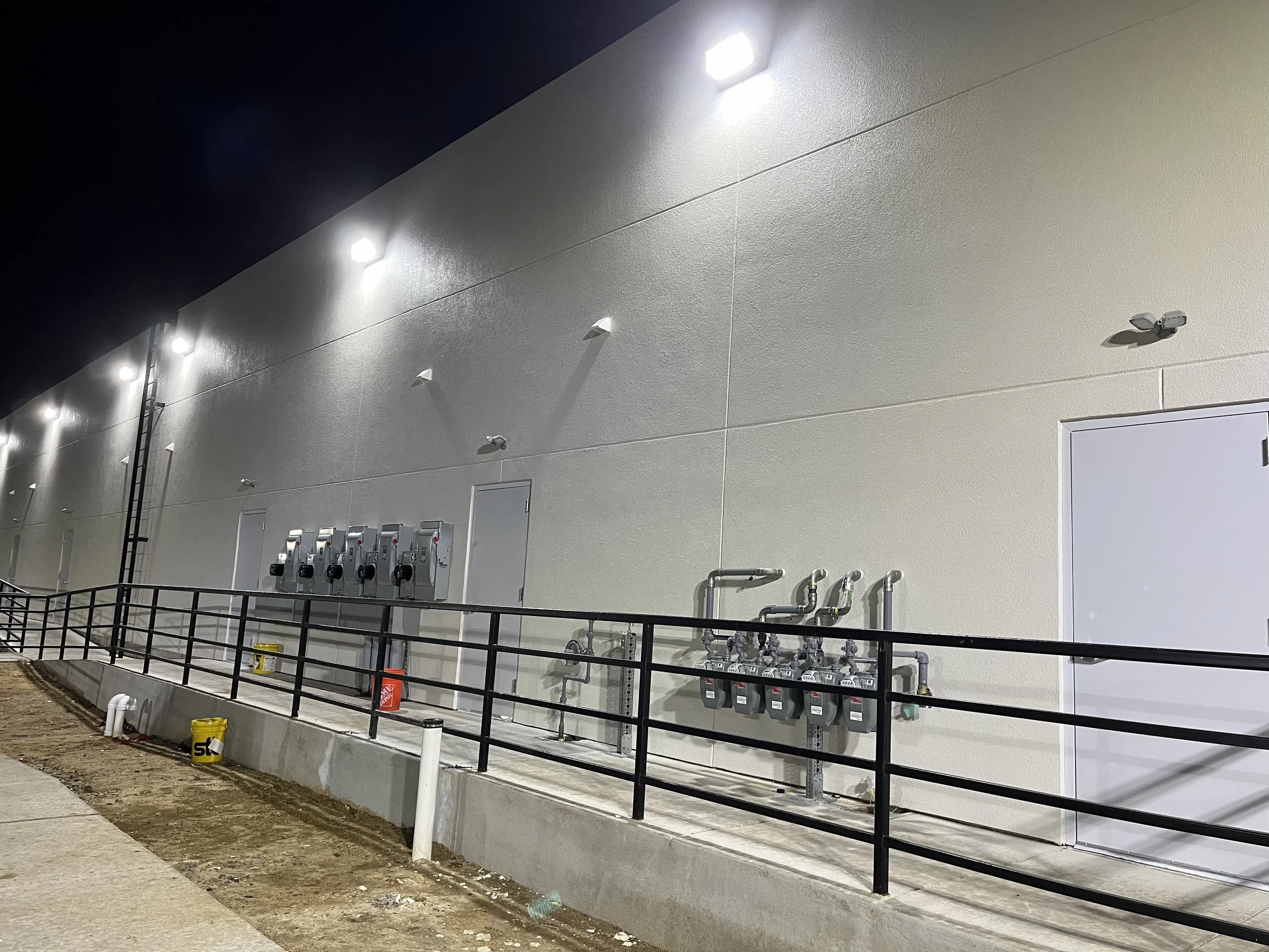 Exterior wall of a building with illumination lights, pipes, and utility meters mounted on it, situated near a sidewalk and a black metal railing.