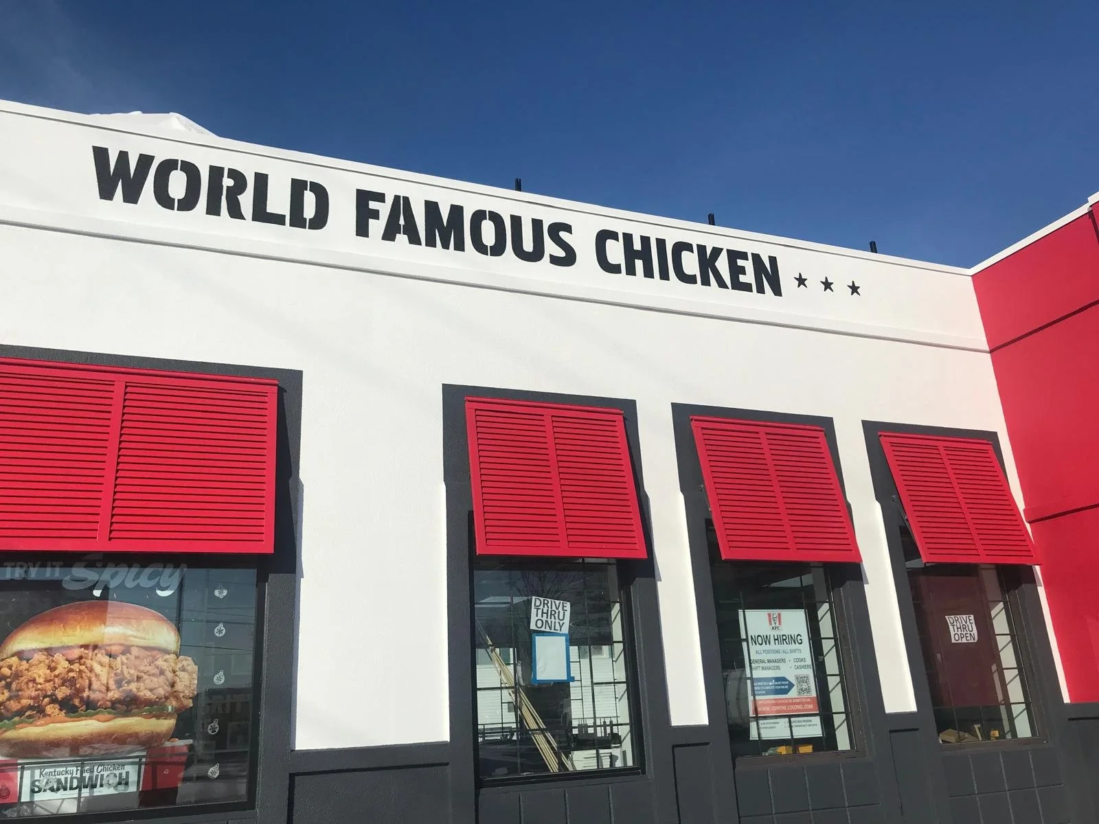 Exterior of a fast-food restaurant with a sign that reads 'World Famous Chicken', red window shutters, and posters advertising chicken sandwiches and drive-thru service.