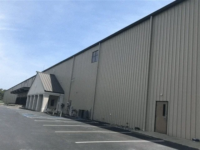Exterior view of a large, beige industrial building with a metal roof and a small entrance area, adjacent to a parking lot with white parking lines.