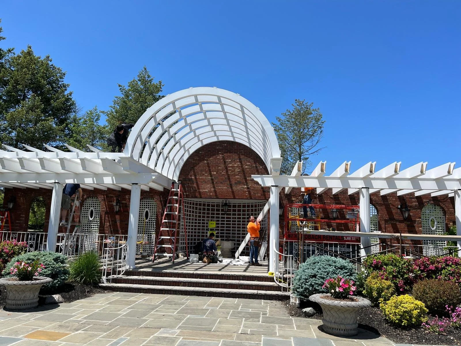 People working on a white pergola structure in front of a brick building with arched windows, surrounded by colorful flowers and green trees under a clear blue sky.