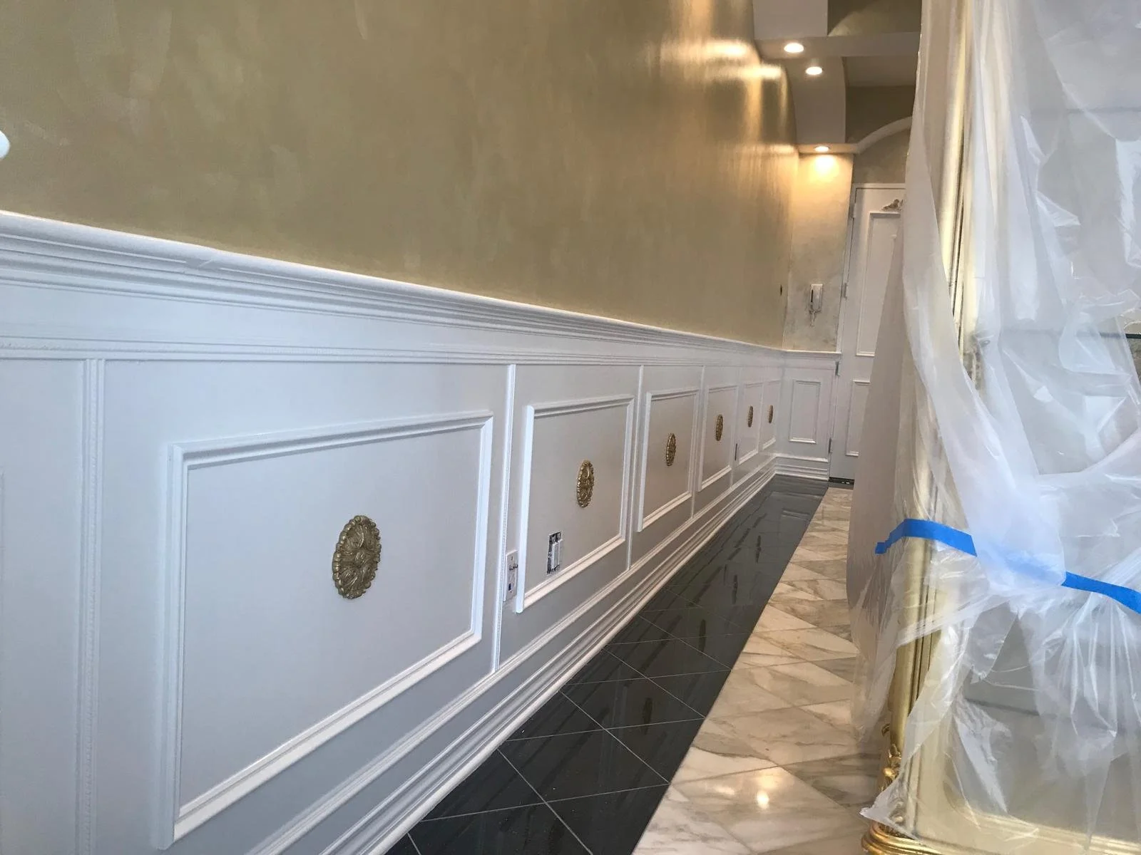 Interior of a room under renovation featuring a white wainscot wall with gold decorative medallions, beige upper wall, black and marble flooring, and plastic cover on furniture.
