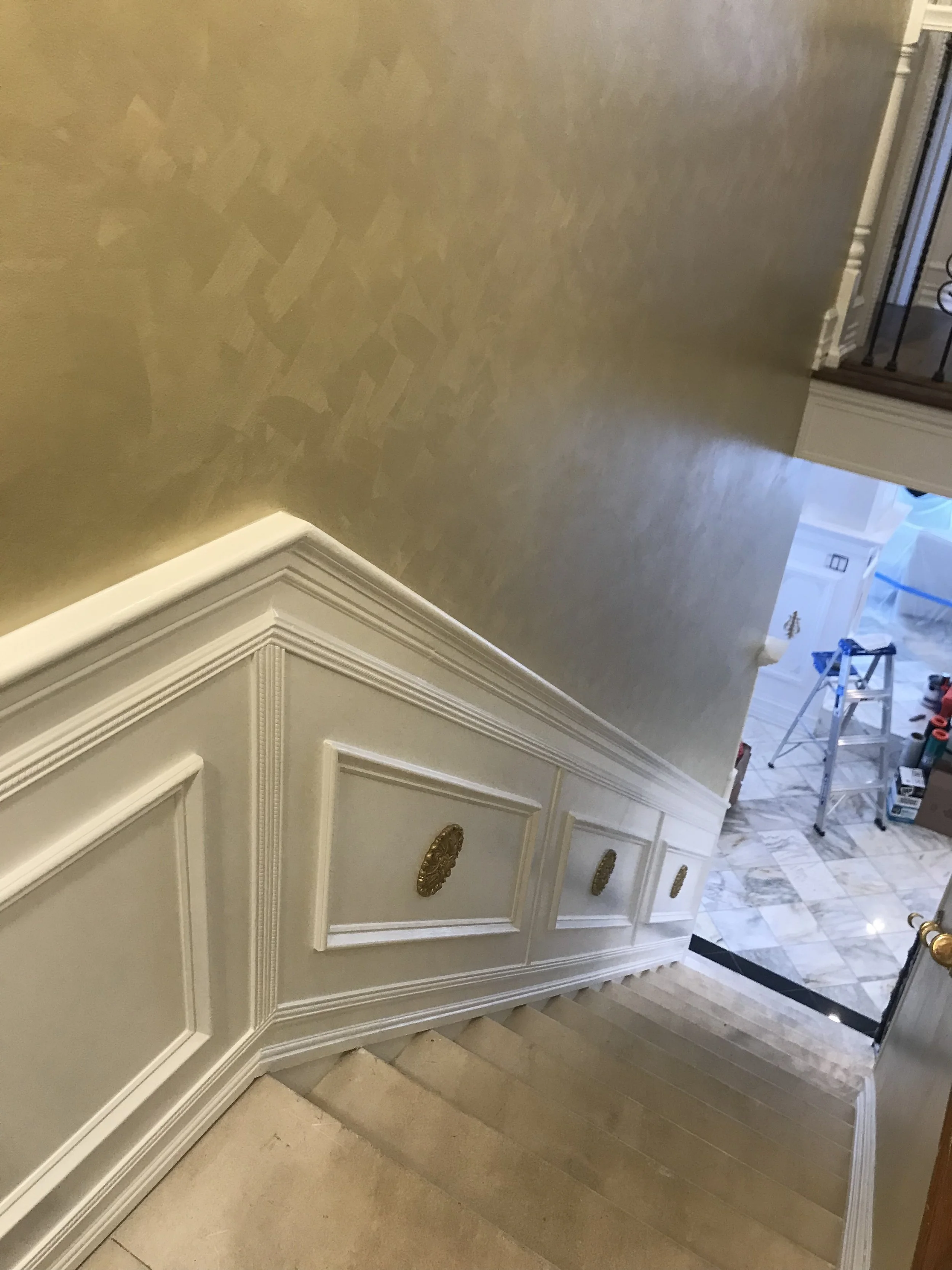Interior view of a staircase with white paneling and gold decorative handles on the cabinet, leading up to a landing with beige and white tiled flooring, and a wall partially painted in a light color.