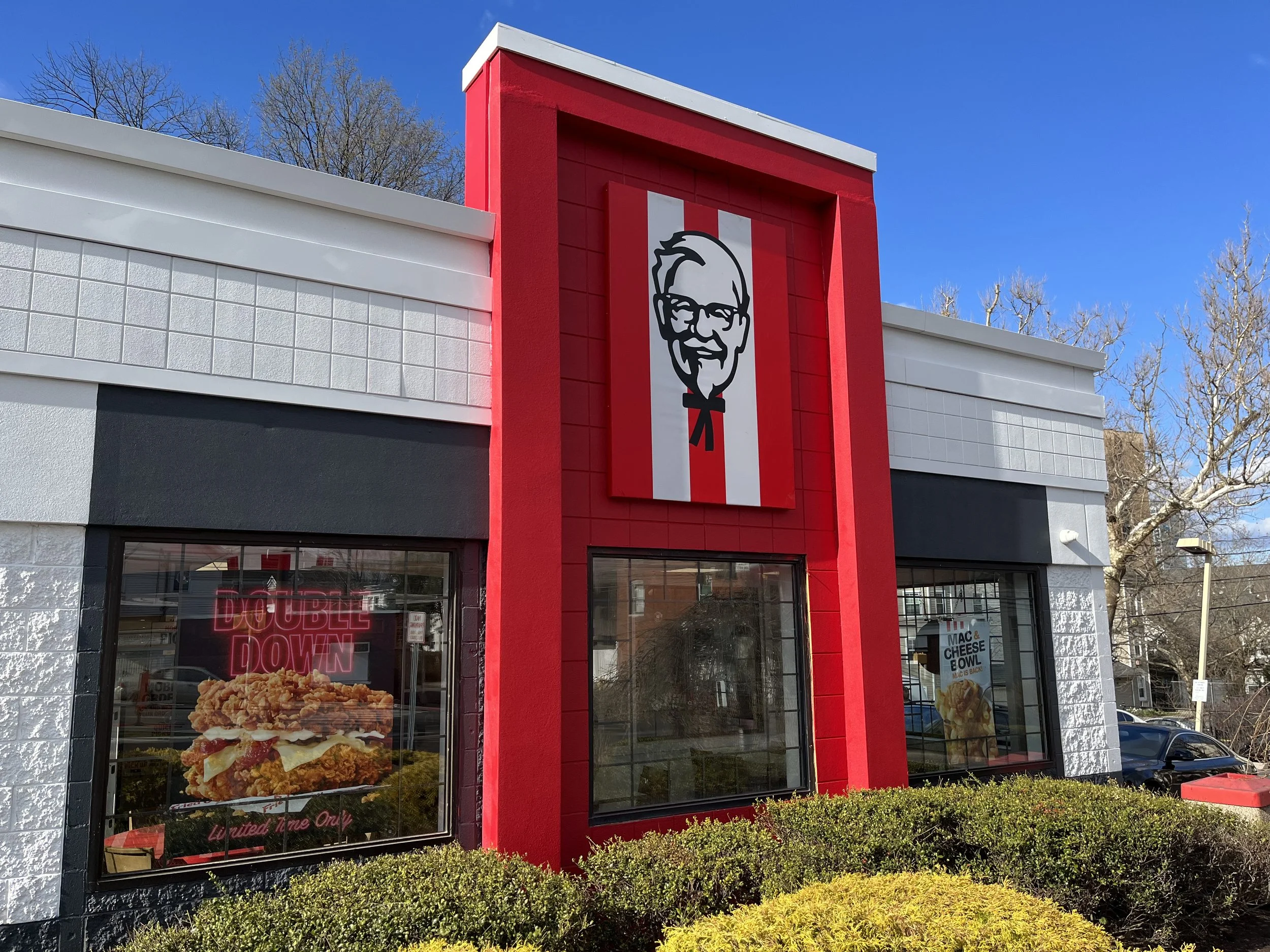 KFC fast food restaurant with a large red vertical sign featuring Colonel Sanders' logo, windows displaying fried chicken, and posters advertising menu items.