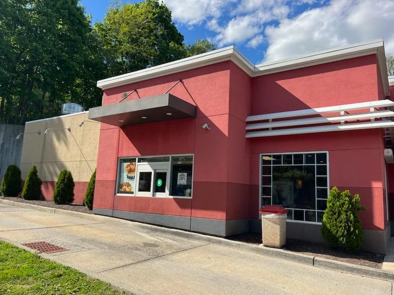 Exterior of a fast-food restaurant with a pink facade, large front window displaying menu items, and a side entrance with a window. Small bushes line the sidewalk and a trash bin is outside.