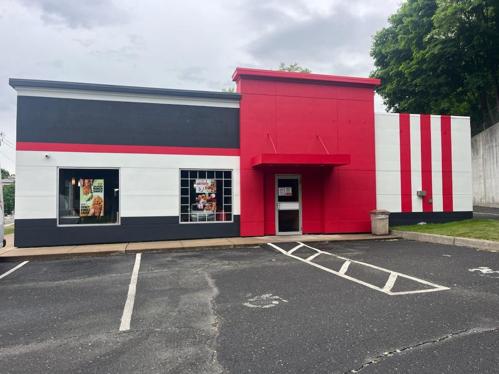 A small fast food restaurant with black, white, and red exterior. There are two windows with posters and a glass door entrance. The parking lot in front has marked spaces, including a handicap spot.