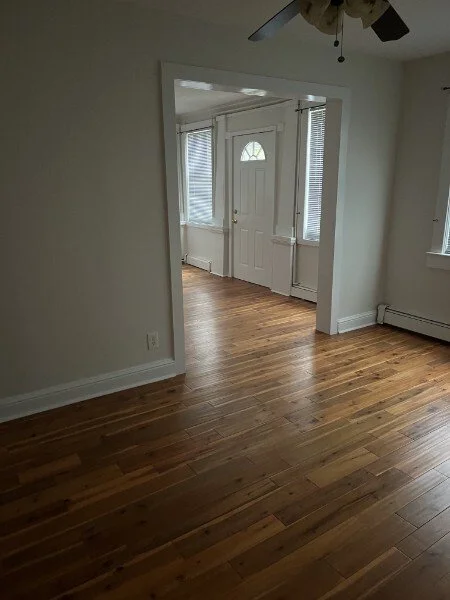 Empty living room area with hardwood floors, white walls, and a ceiling fan, leading to a front door with windows.