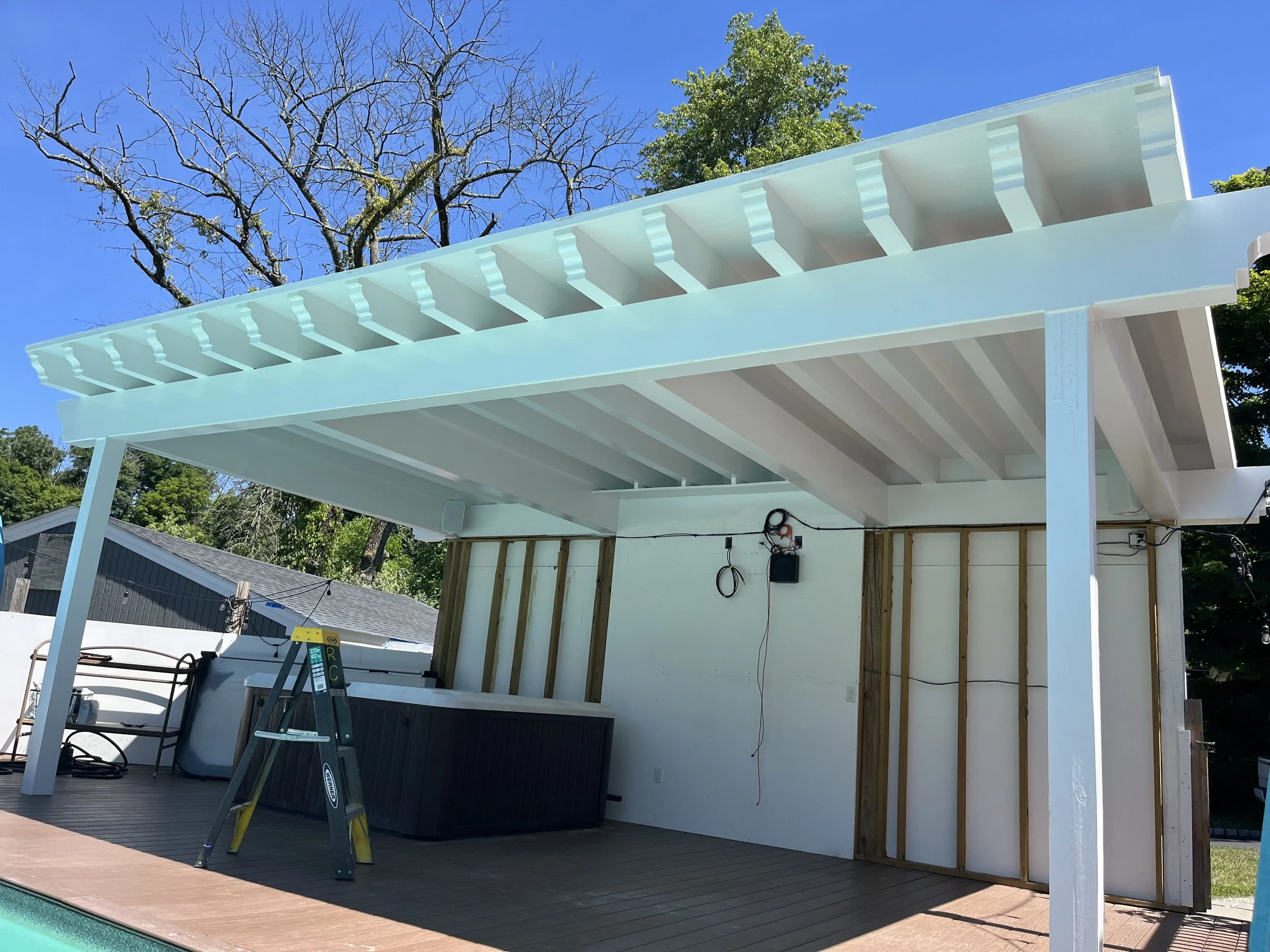 Construction site showing an outdoor patio or deck area with a partially built white pergola or roof structure, some electrical wiring, a ladder, outdoor furniture including a hot tub, and a swimming pool in the foreground. A tree with bare branches 