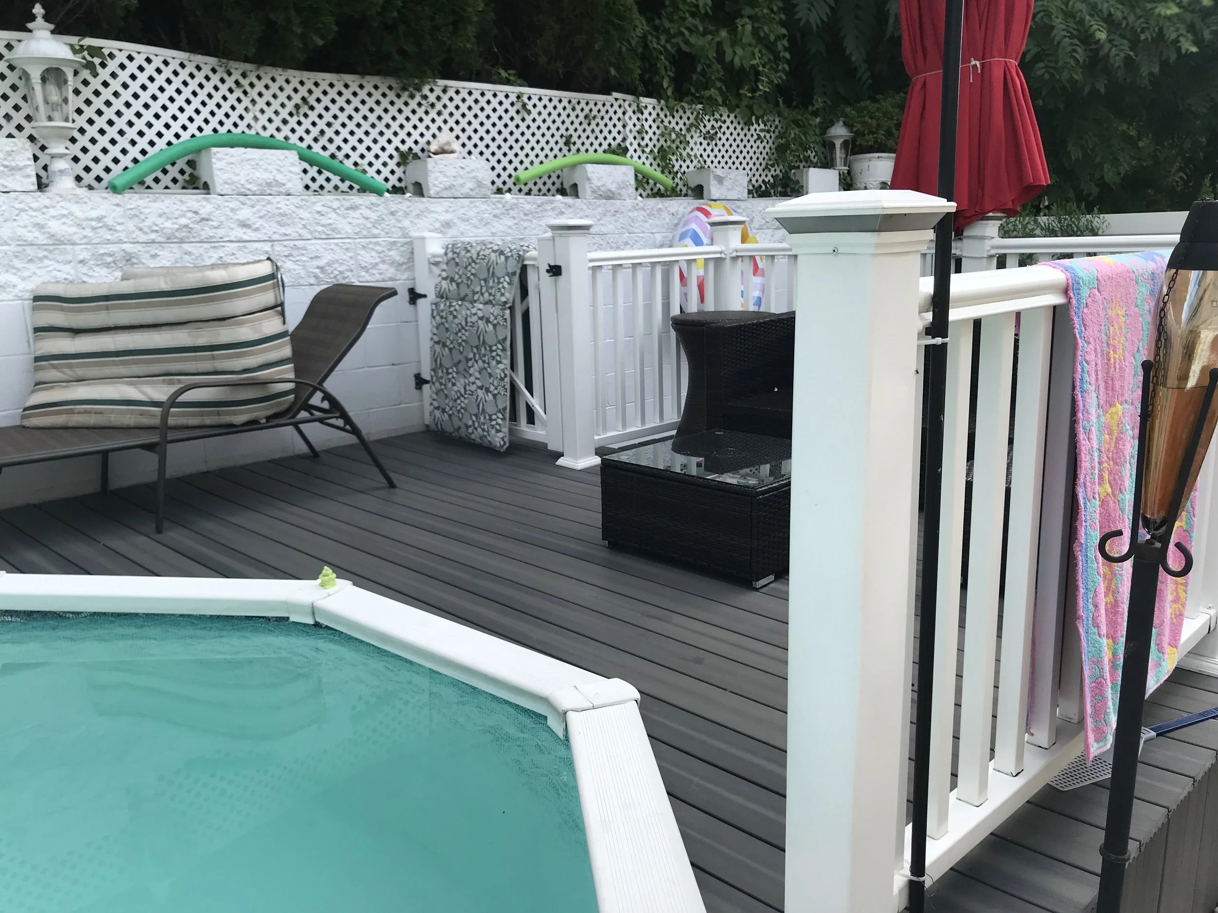 Residential backyard pool area featuring a small swimming pool, poolside chairs with striped and dark cushions, towel draped over a railing, a closed red umbrella, decorative fencing, and lush green trees in the background.