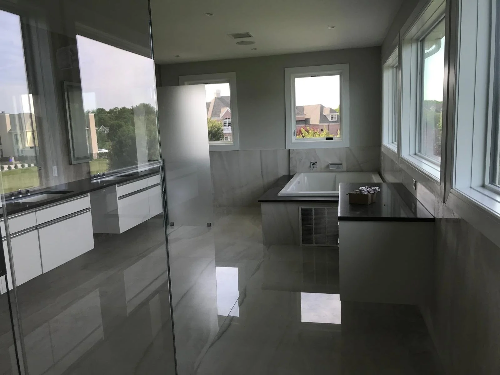 Modern bathroom with large windows, bathtub, marble tiles, and black countertops, seen through a glass partition.