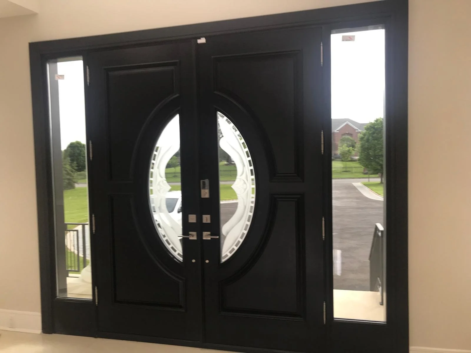 A black front door with oval glass design and two side windows, with a view of a driveway, street, green lawn, trees, and houses outside.