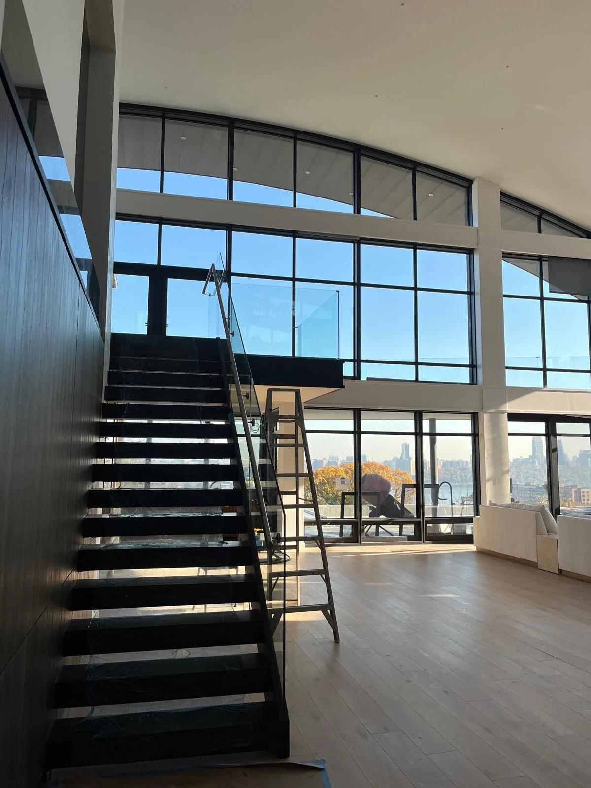 Interior view of a modern building with large glass windows, a staircase, and cityscape in the background.