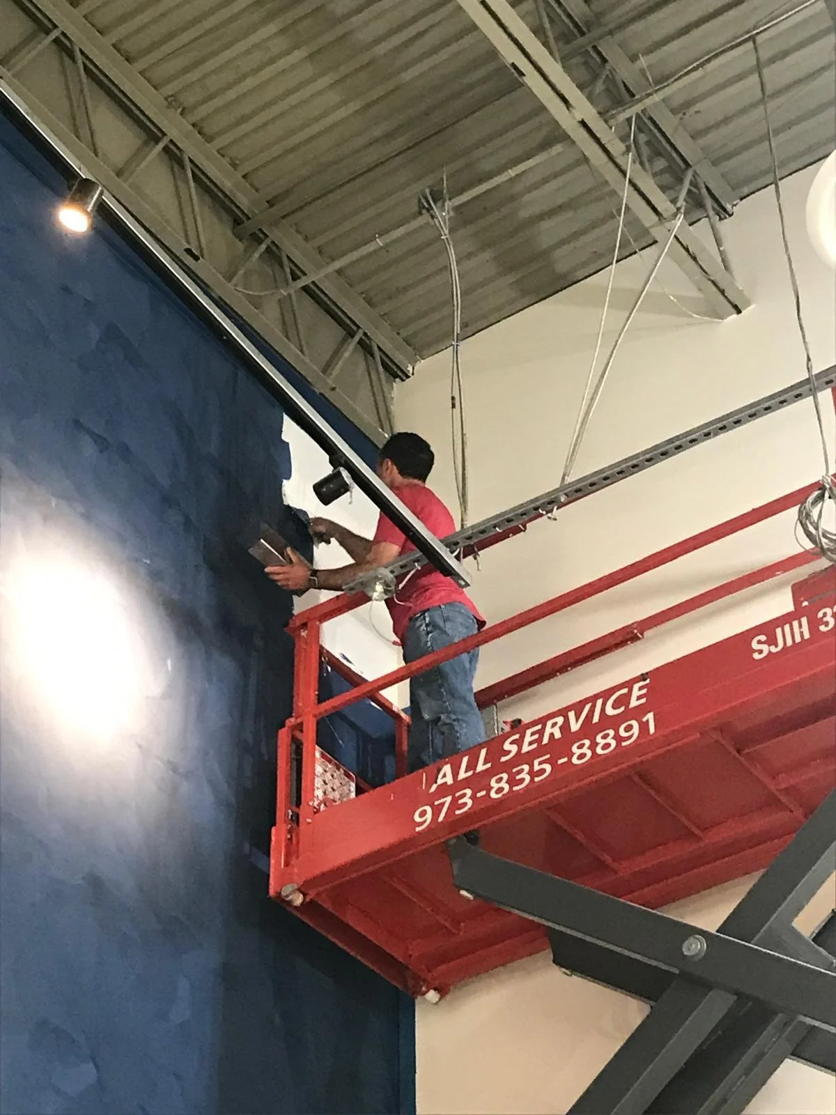 Two workers on a red lift working on a large blue wall display in an indoor setting.