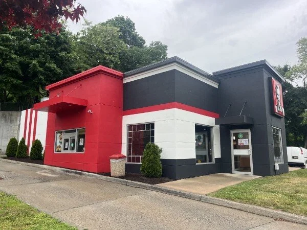 A Taco Bell fast-food restaurant with red, black, and white exterior walls, a small front window for placing orders, and a drive-thru window. There are some bushes and trees nearby, and a parking lot with a few cars.