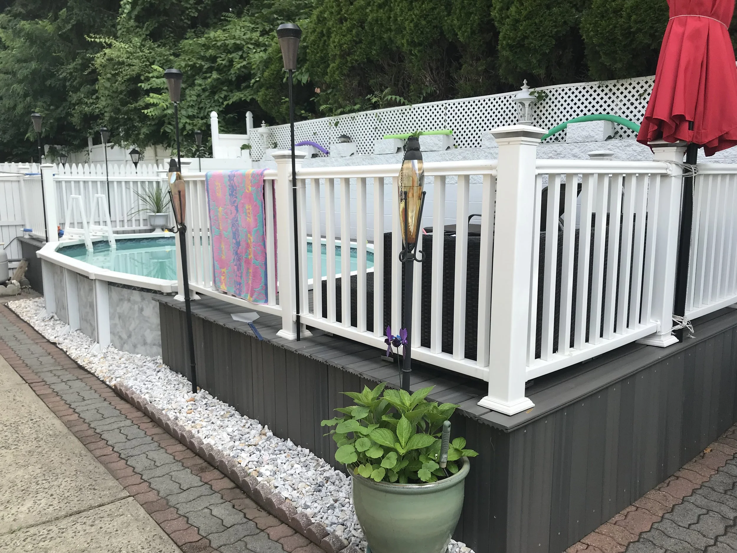 A backyard with a white fence, a hot tub, potted plants, a colorful towel hanging, and a red patio umbrella.