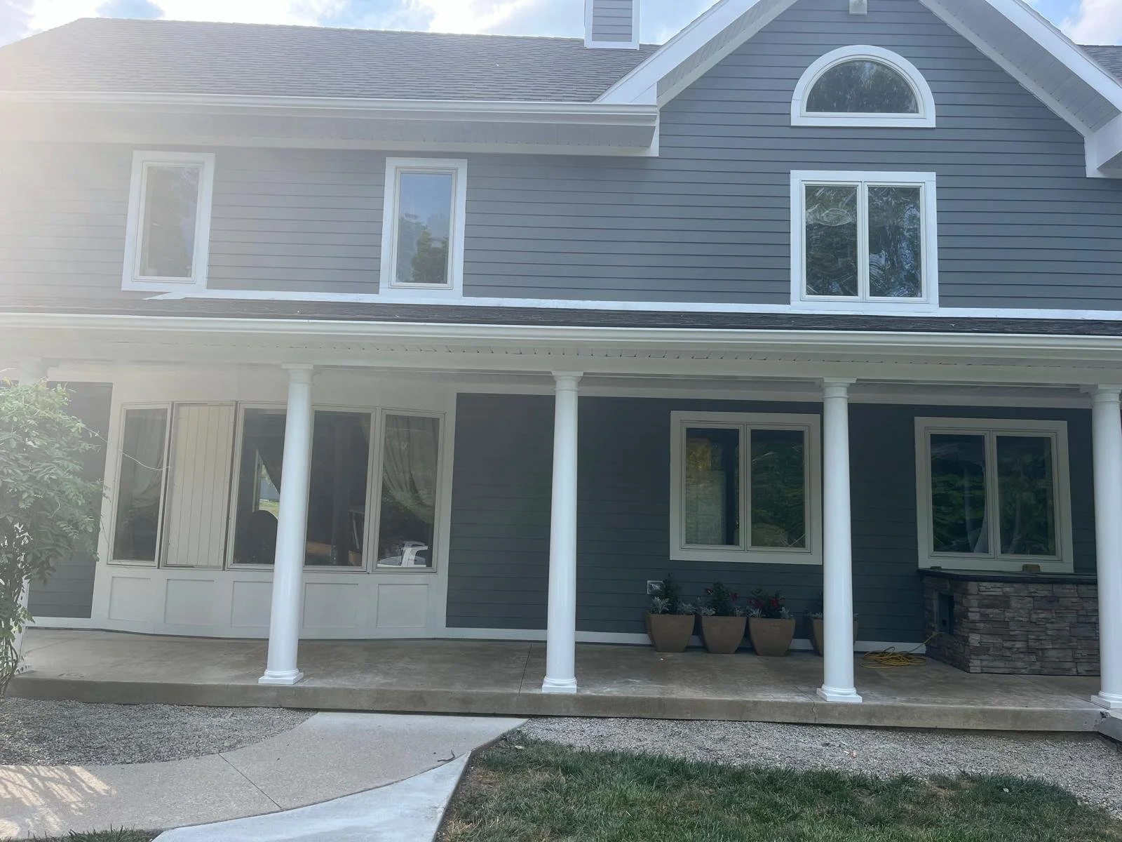 Front view of a gray two-story house with a covered porch supported by white columns, multiple white-framed windows, and a small garden with potted plants