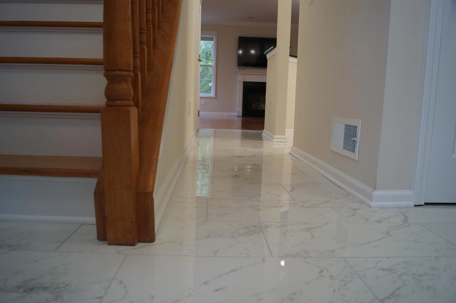 View of a hallway with shiny marble floors, a staircase with wooden railing on the left, beige walls, and a living room with a fireplace and TV in the background.