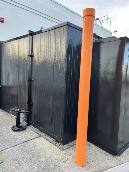 Black metal utility enclosure with an orange safety pipe in front and a concrete sidewalk.