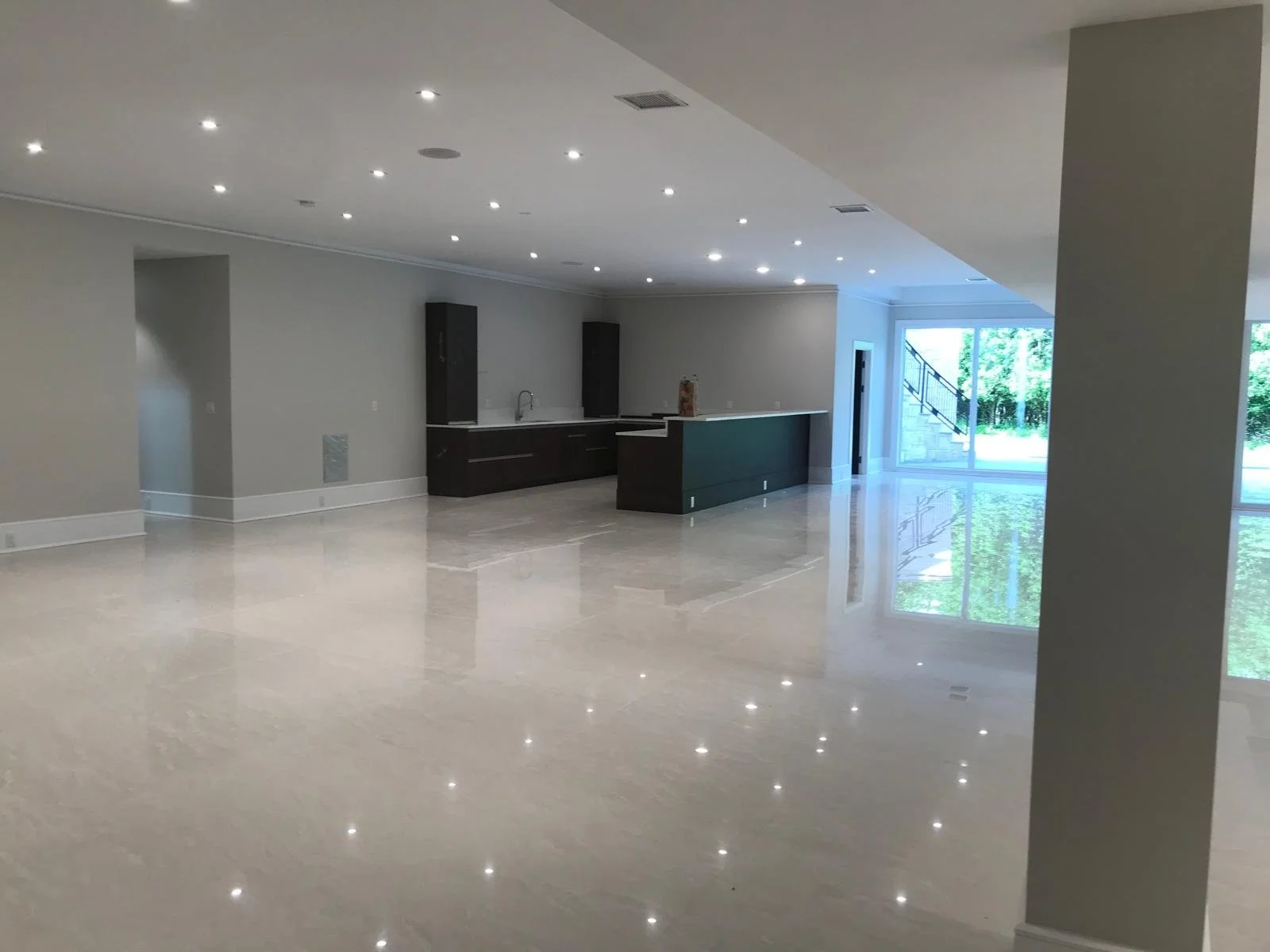 Empty modern open-concept living space with high ceilings, recessed lighting, large beige tile flooring, a kitchen with dark cabinetry, a white countertop, and a green island, with sliding glass doors leading outside to a lush green yard.