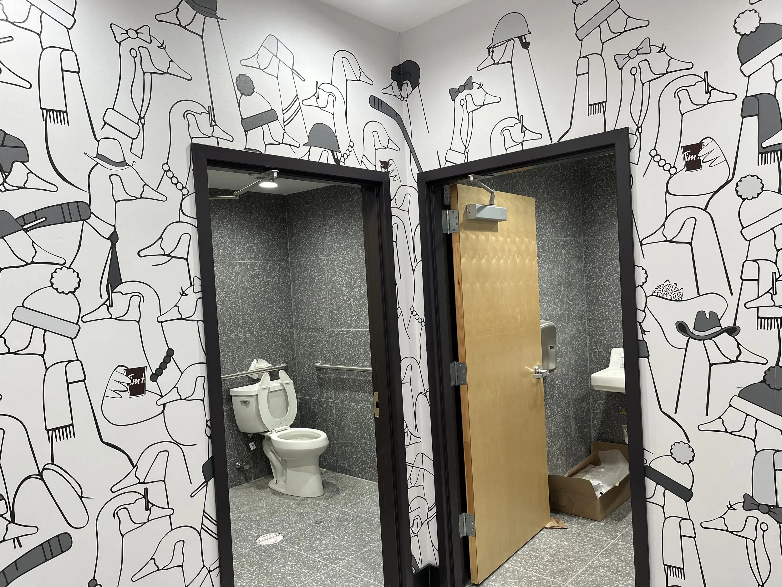Public restroom with two stalls. One stall door is open, revealing a white toilet inside. The walls have black and white abstract line art illustrations of various characters, including people wearing hats and holding drinks.