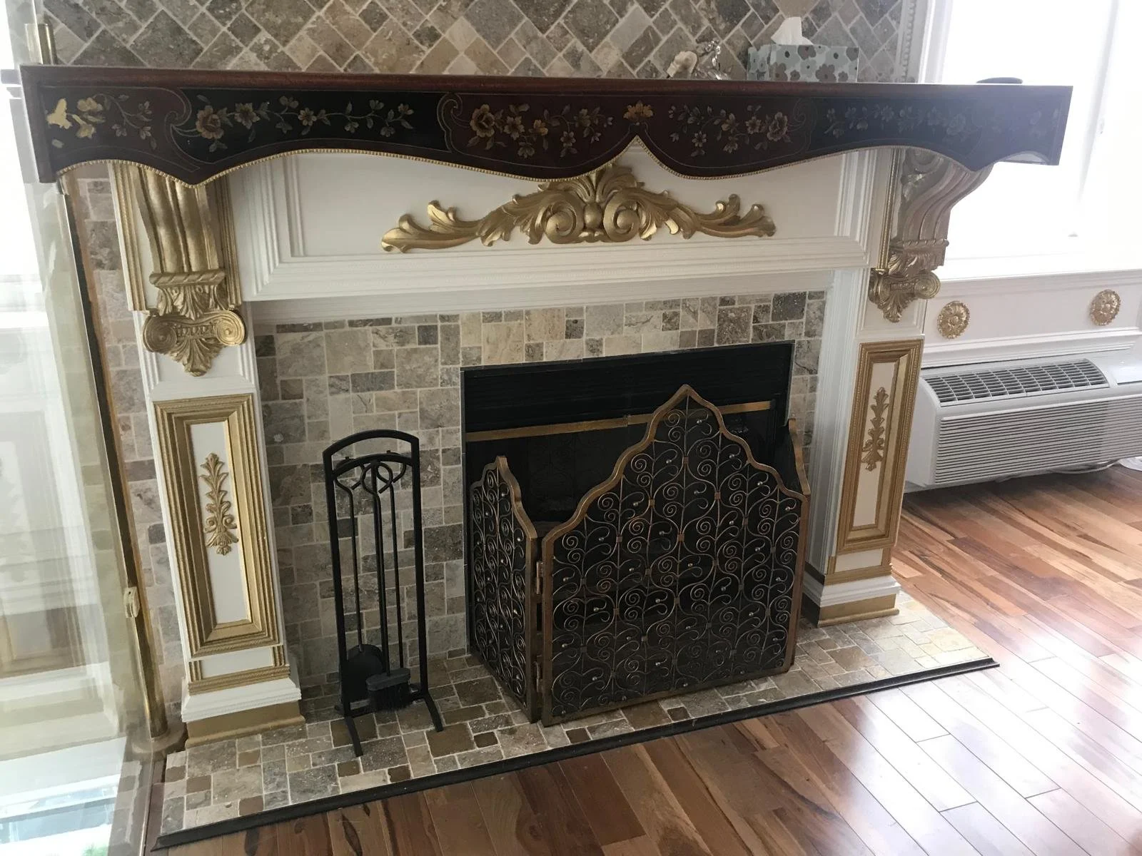 Elegant fireplace with ornate gold and white molding, black iron fire screen with gold accents, and decorative tools. The fireplace has a tiled surround and adjacent to an air conditioning unit, with hardwood flooring in the room.