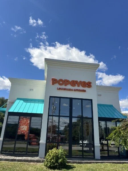 Popeyes Louisiana Kitchen fast food restaurant with white exterior, large glass windows, blue awnings, and a sign reading "Popeyes Louisiana Kitchen" under a partly cloudy blue sky.