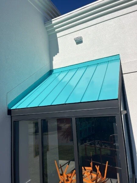 A photo of a light blue metal awning above glass sliding doors on a stucco building, with an outdoor table and chairs inside visible through the glass.