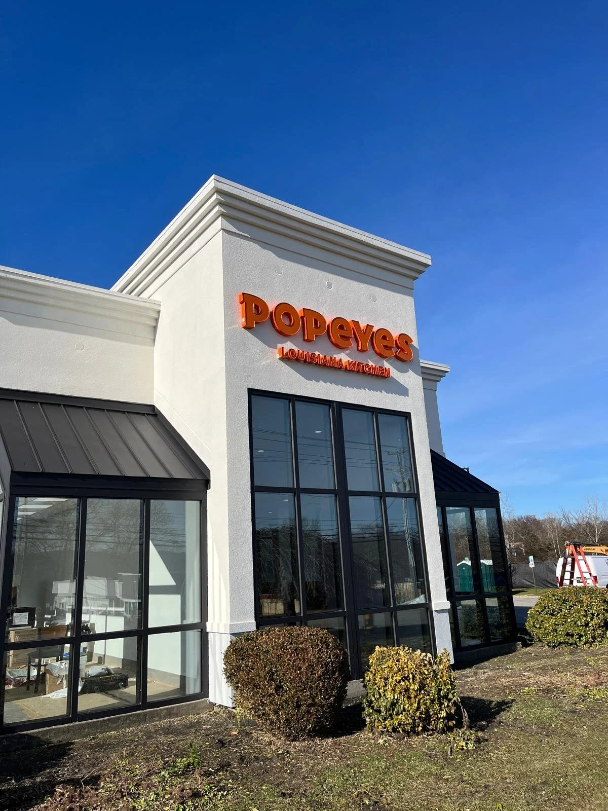 Exterior of Popeyes Louisiana Kitchen restaurant with large windows, white walls, and red signage, under a clear blue sky.