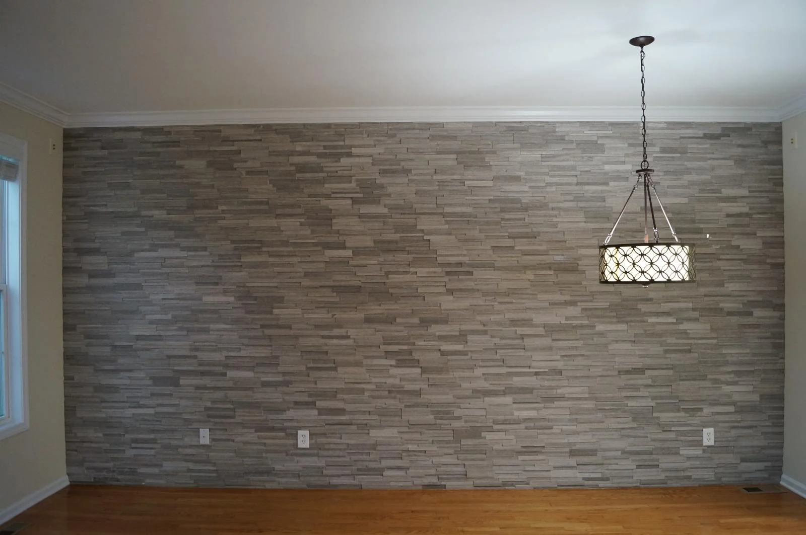 Room with a grey stone accent wall, chandelier hanging from the ceiling, hardwood flooring, and a window on the left with blinds.