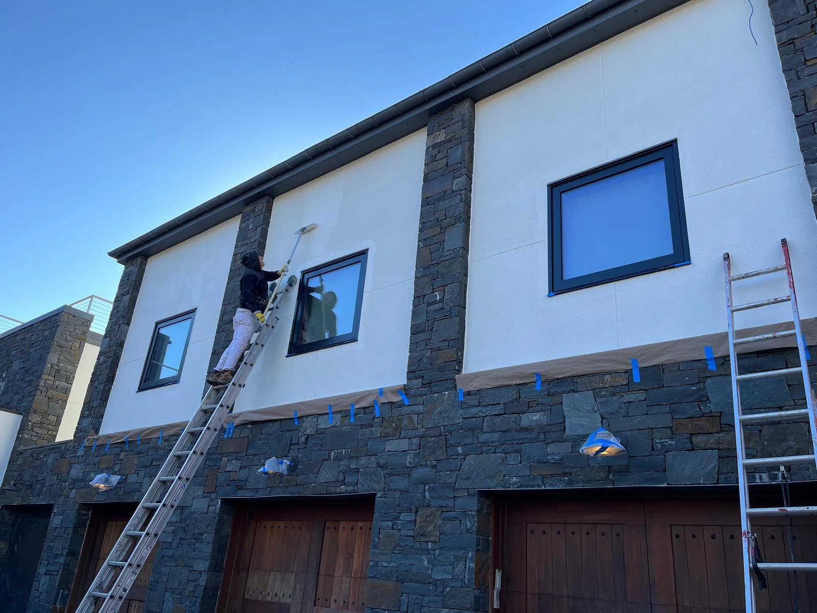 Worker on a ladder painting the exterior wall of a modern house with white and stone features, under a clear blue sky.