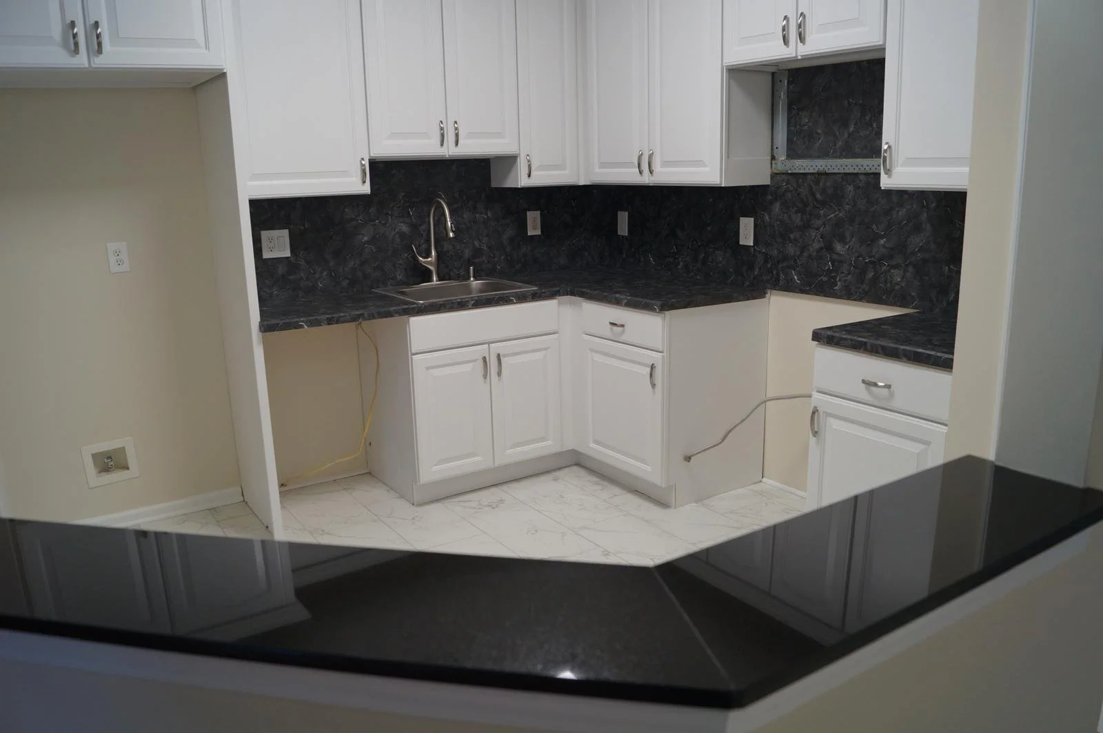 Kitchen with white cabinets, black marble countertops, and a stainless steel sink. There are multiple electrical outlets on the walls, and some cabinets are missing doors, showing the interior shelving. The floor is white marble with gray veining.