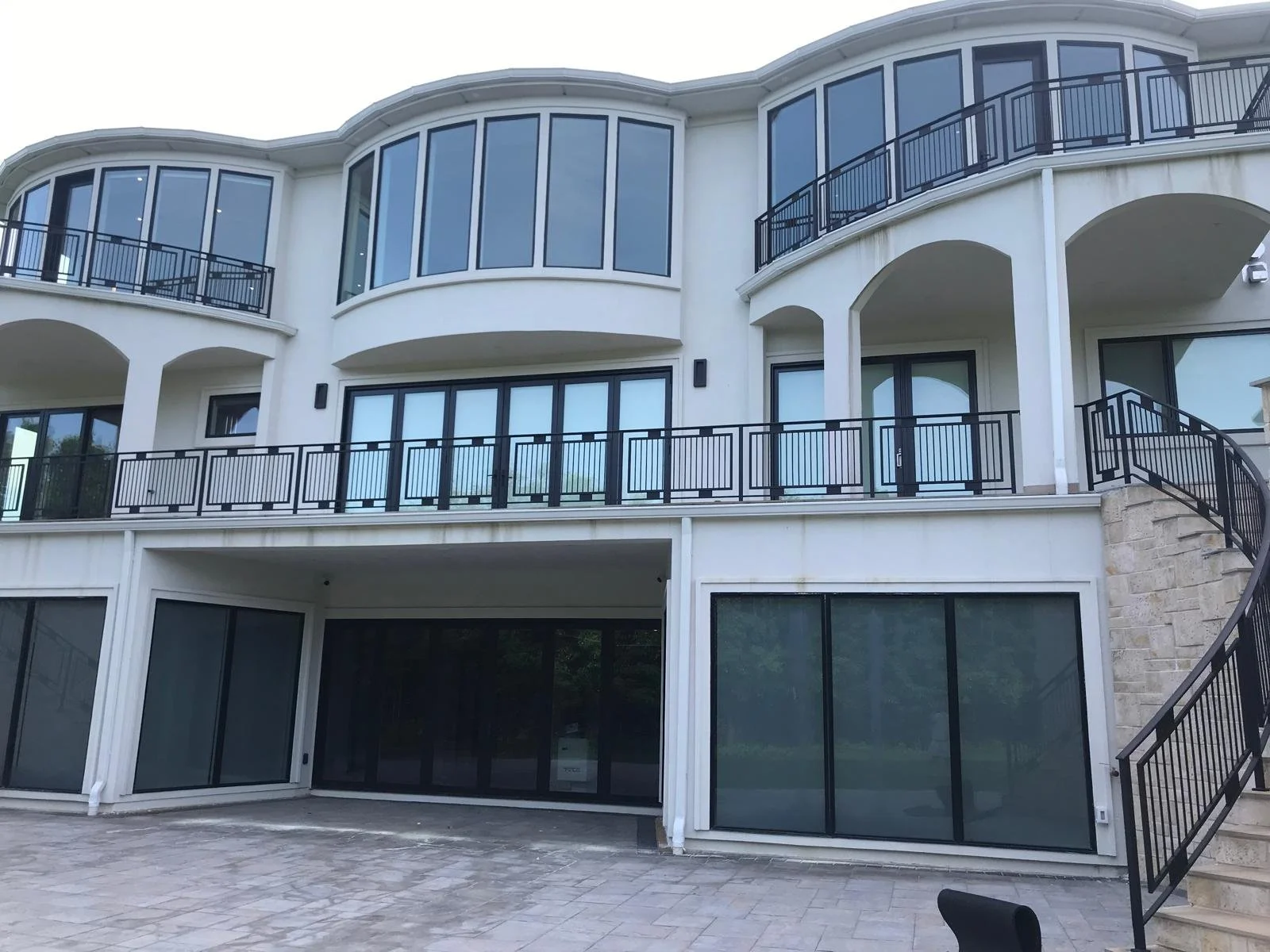 Multi-story modern residential building with large windows, glass doors, and black metal railings on the balconies, with a stone and brick exterior and a paved outdoor area.