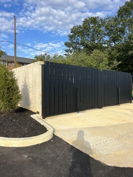 Black wooden gate with a beige concrete wall to its left, a curved parking curb, and a concrete driveway under a partly cloudy sky with trees in the background.