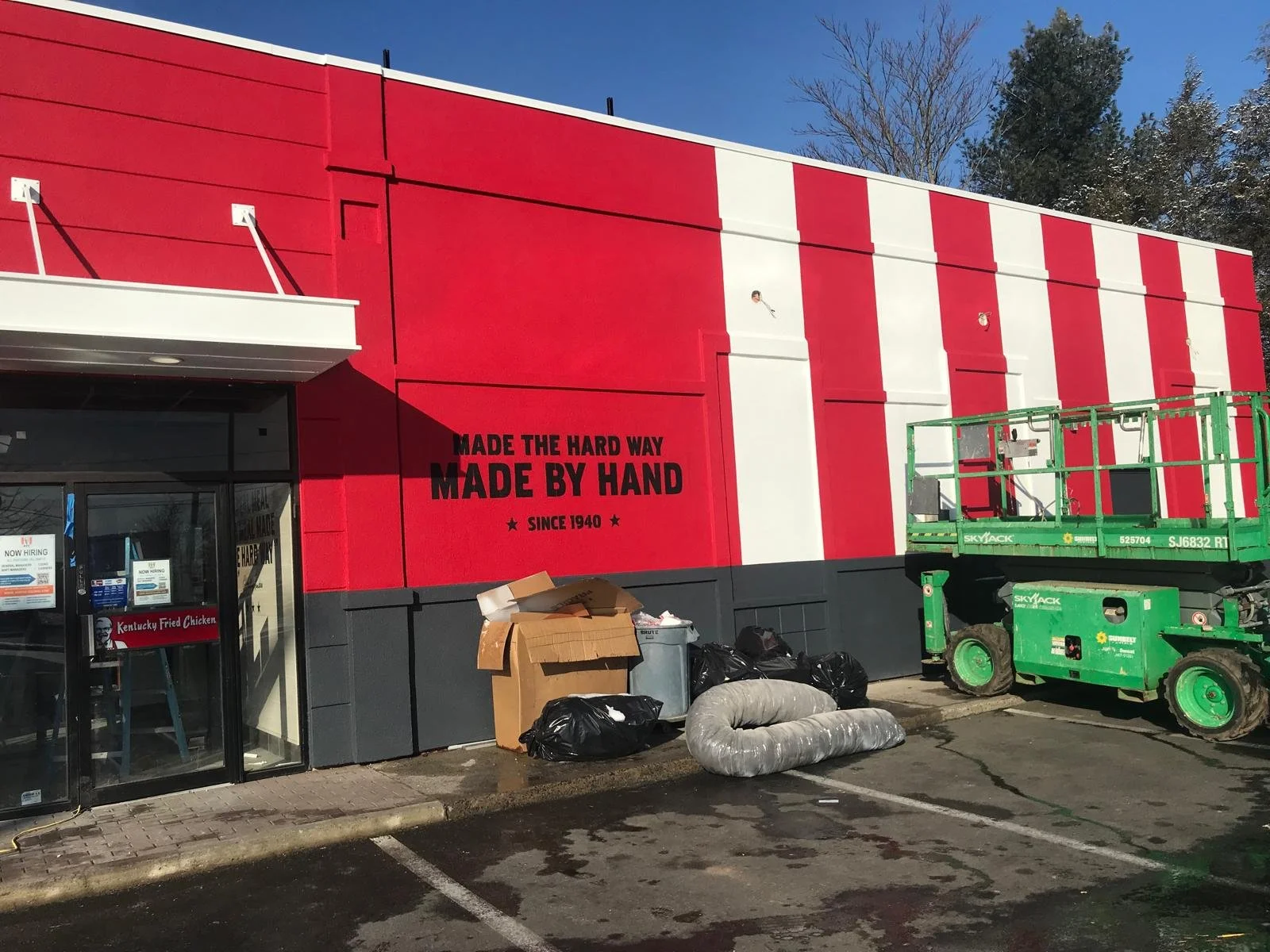 Exterior of a Kentucky Fried Chicken restaurant with a bright red striped facade, a black lower section, and a glass entrance door. There are some trash bags, cardboard, and a flexible duct on the ground outside, along with a green scissor lift parke